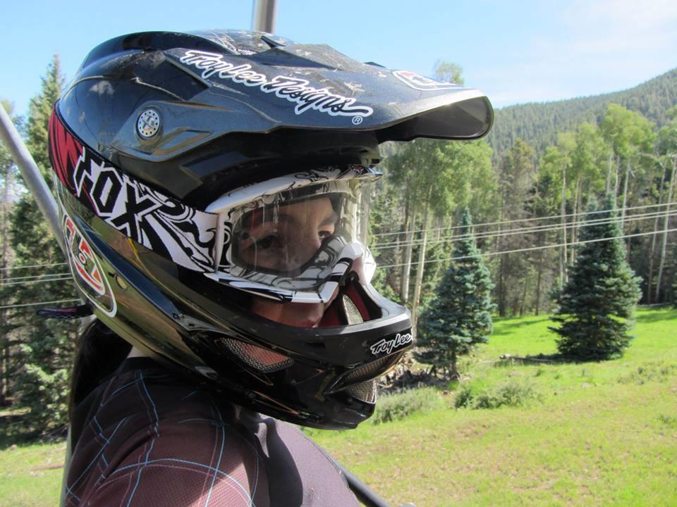 A person wearing a black and white motocross helmet sits on a ski lift, with a lush green landscape and mountains in the background. The image captures the excitement of outdoor sports with trees and power lines visible behind them. Angel Fire Bike Park mountain bike trail.