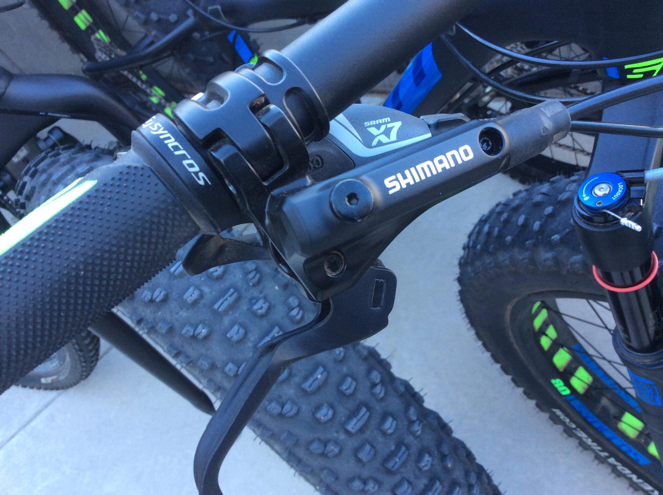 Close-up view of a mountain bike handlebar featuring a Shimano brake lever and SRAM X7 gear shifter, with textured grips and a black and green color scheme. The background shows part of another bicycle and the textured surface of fat tires.