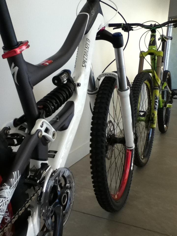 Two mountain bikes are parked side by side against a plain wall. The bike in the foreground is predominantly white with a sleek design, featuring a prominent suspension system and red accents on the rims. The second bike, partially visible in the background, is green with black components and knobby tires, suggesting it is built for rugged terrain. The indoor setting has a smooth concrete floor, emphasizing the bikes' sporty appearance.