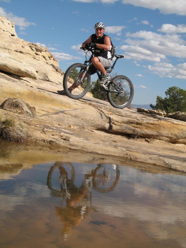A cyclist poses on a rocky terrain next to a small puddle, with their reflection visible in the water. The scene is set under a blue sky with scattered clouds, showcasing a landscape of natural rock formations and sparse vegetation.