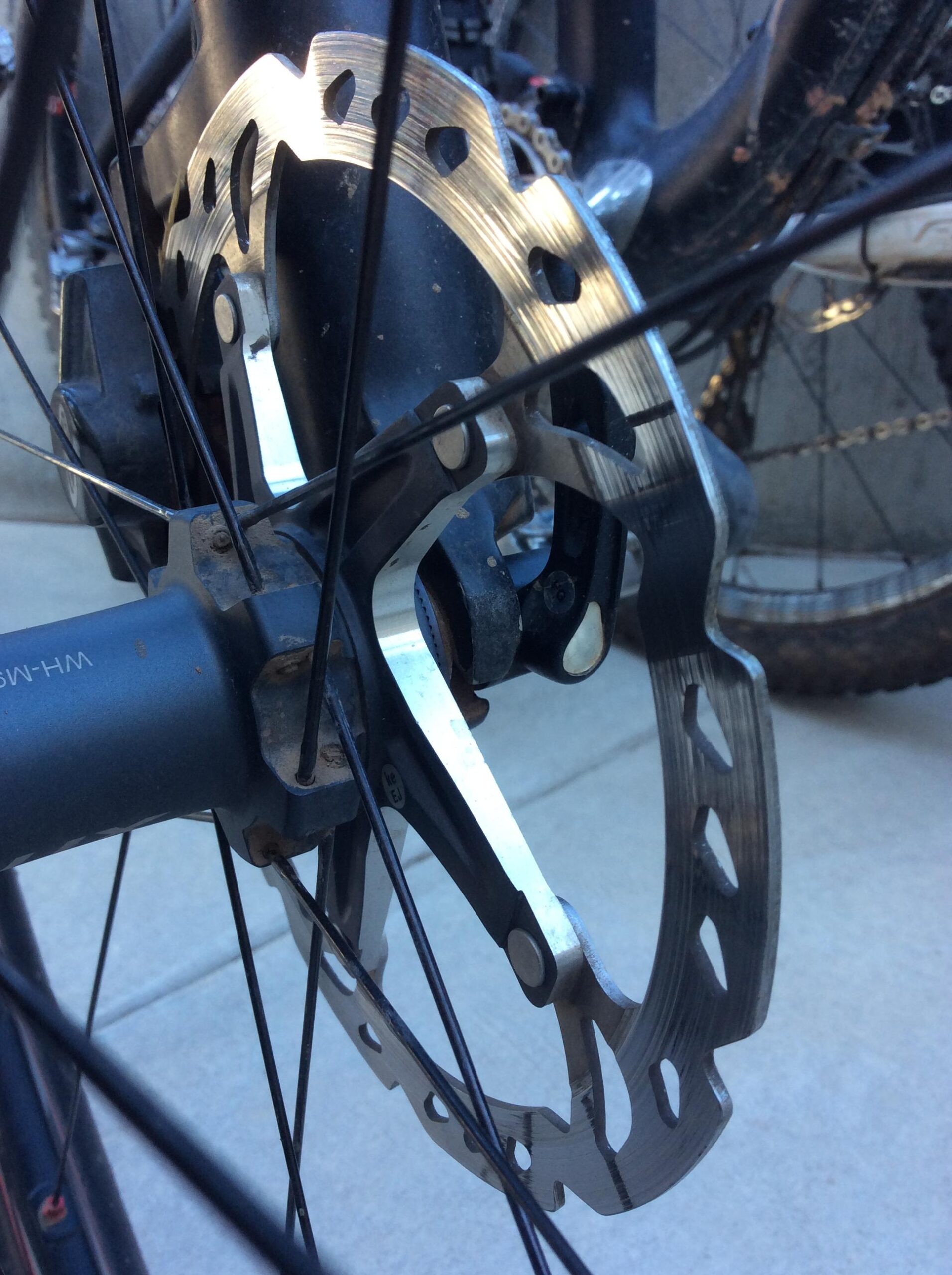 Close-up image of a bicycle's disc brake system, showing the metallic brake rotor, part of the hub assembly, and bicycle spokes. The background features another bicycle wheel and a concrete surface.