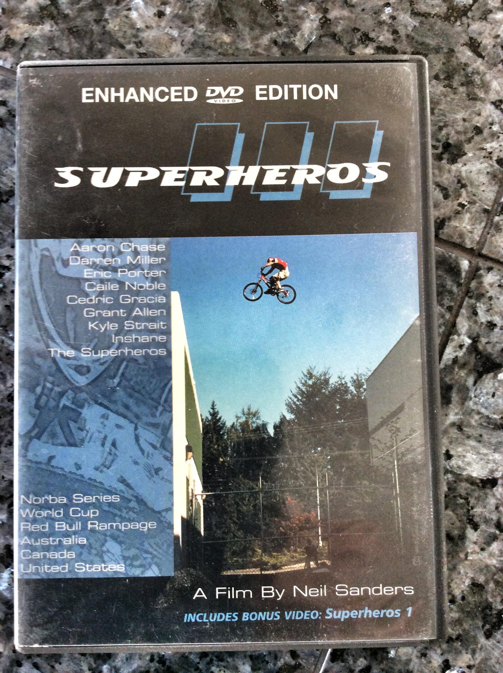 A DVD cover titled "Superheros," featuring a BMX rider performing a jump in a blue sky. The cover lists various riders, including Aaron Chase and Eric Porter, and mentions it is an enhanced edition with a bonus video. Additional text highlights events such as the Norba Series and Red Bull Rampage. The film is directed by Neil Sanders. The background features trees and buildings, suggesting an outdoor setting.