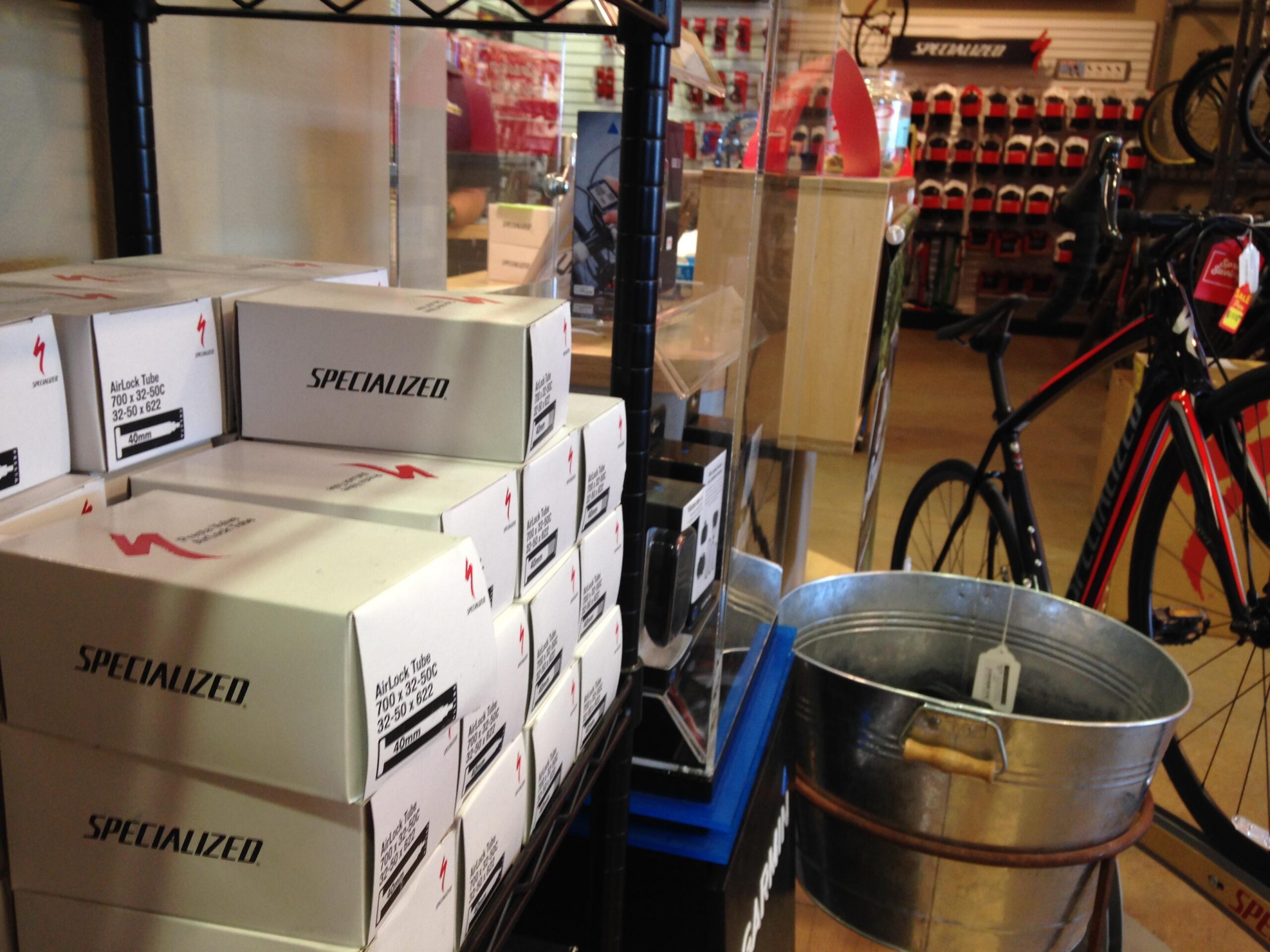 A display in a bicycle shop featuring boxes of Specialized airlock tubes on a shelf, with a metal bucket in the foreground and a black bicycle in the background. The shop interior includes various bike accessories and gear displayed on the walls.