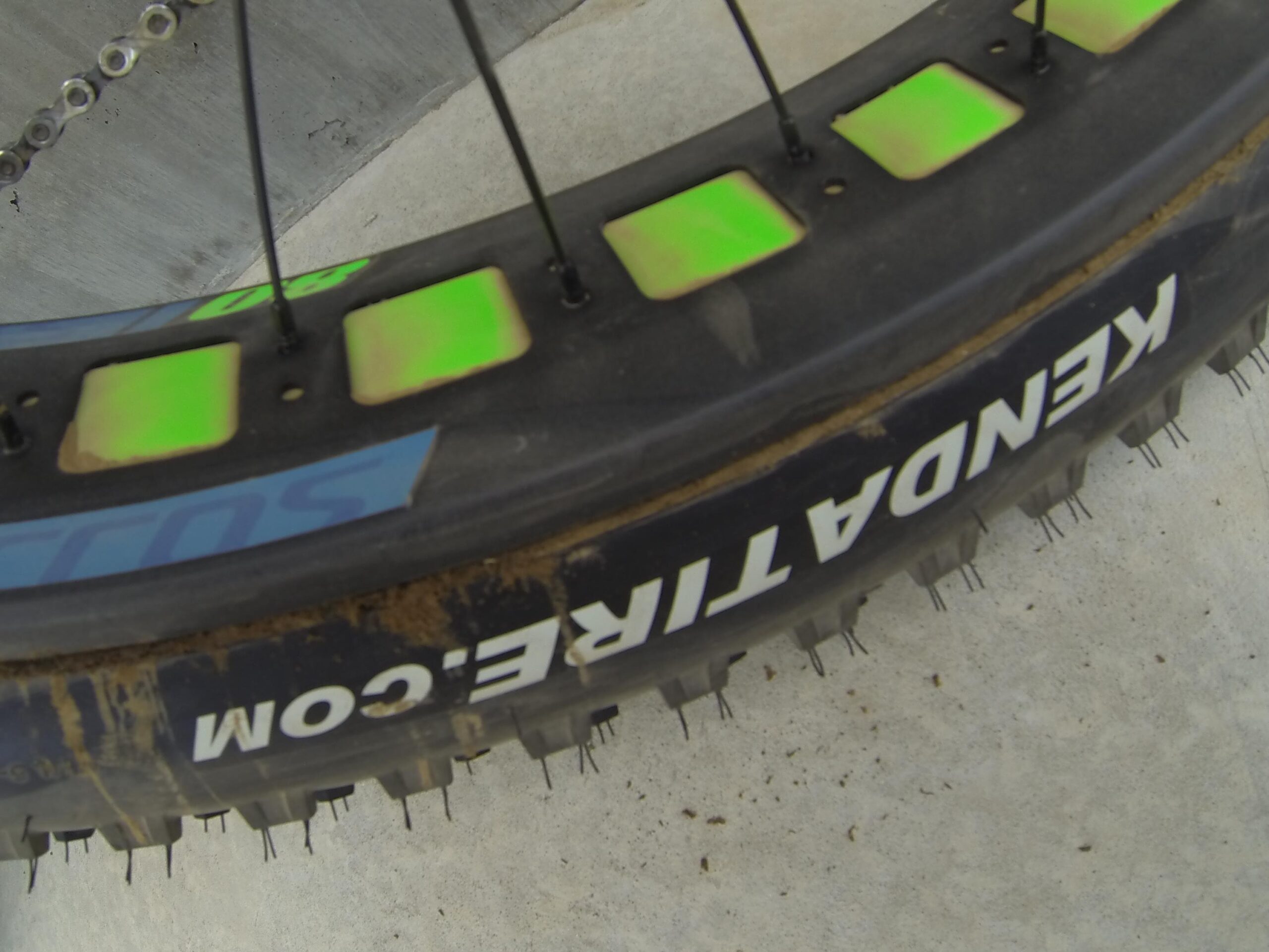 Scott Big Ed: Close-up view of a bicycle tire showcasing the tread pattern, with visible mud on the sidewall. The tire features neon green accents and branding reading "Kenda Tires" along the rim. The background is a concrete surface with some chain links visible above the tire.