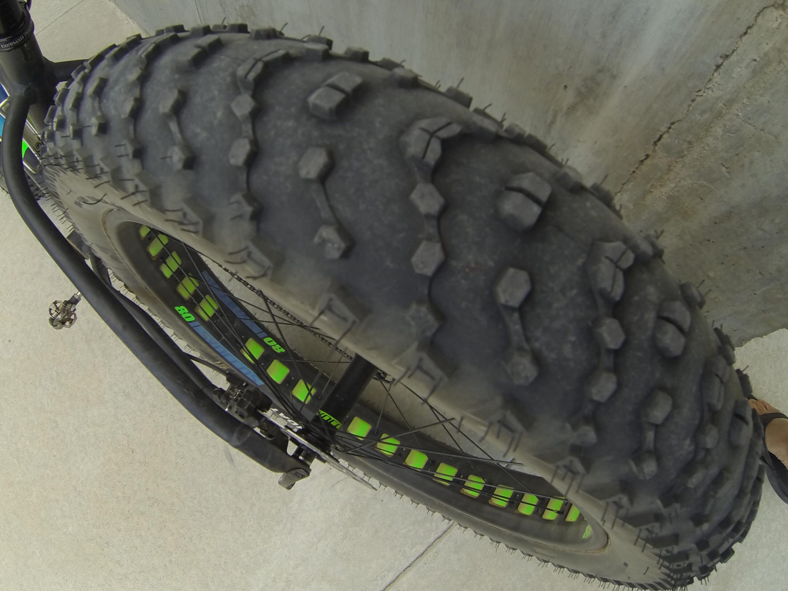Scott Big Ed: Close-up view of a fat bike tire with a rugged tread pattern. The tire shows signs of wear and dirt, and is positioned against a concrete surface. The bike's spokes and part of the pedal are also visible, along with a foot wearing a sandal in the bottom corner of the image.