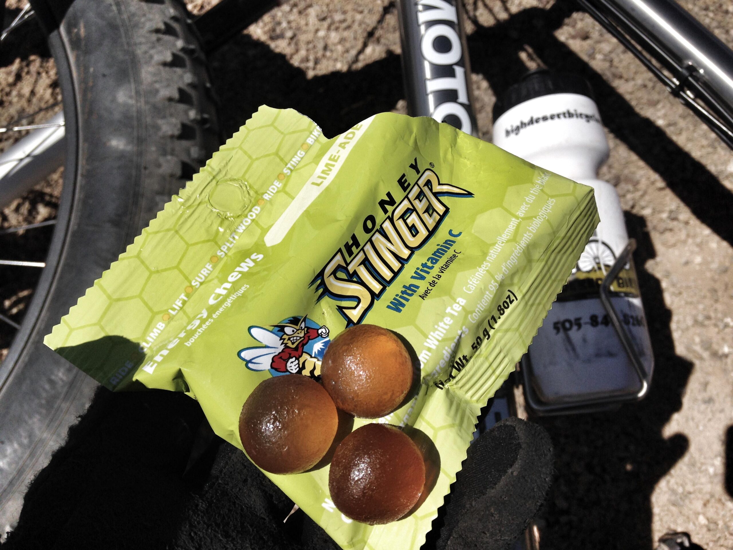 A close-up view of a pack of Honey Stinger energy chews in a lime flavor, displaying three chews in front of the partially torn package. The background features a bike tire and a water bottle, suggesting an outdoor adventure or biking context.