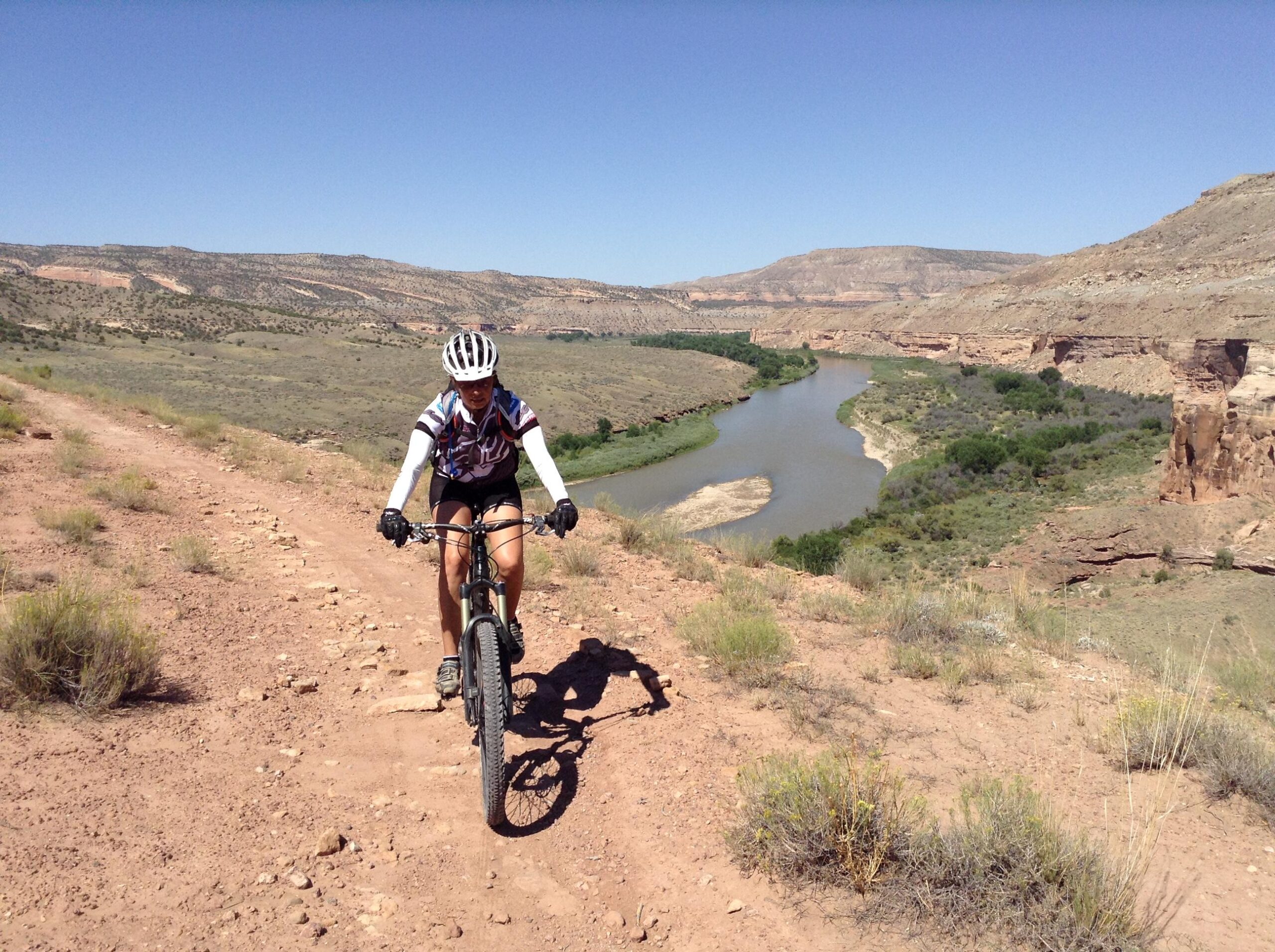 Specialized Stumpjumper FSR Pro: A person riding a mountain bike along a dirt path, with a scenic backdrop of hills and a river curving through a green valley under a bright blue sky.