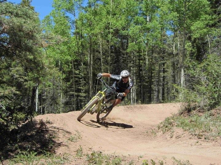 A mountain biker skillfully leans into a turn on a dirt trail surrounded by green trees, showcasing agility and control as he navigates the course. Angel Fire Bike Park mountain bike trail.