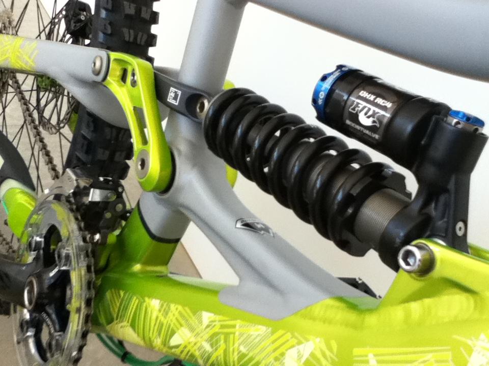 Close-up view of a bicycle's rear suspension, showcasing a coil shock absorber and linkage system. The frame features a vibrant green and gray design, with visible drivetrain components including a chain and crankset.