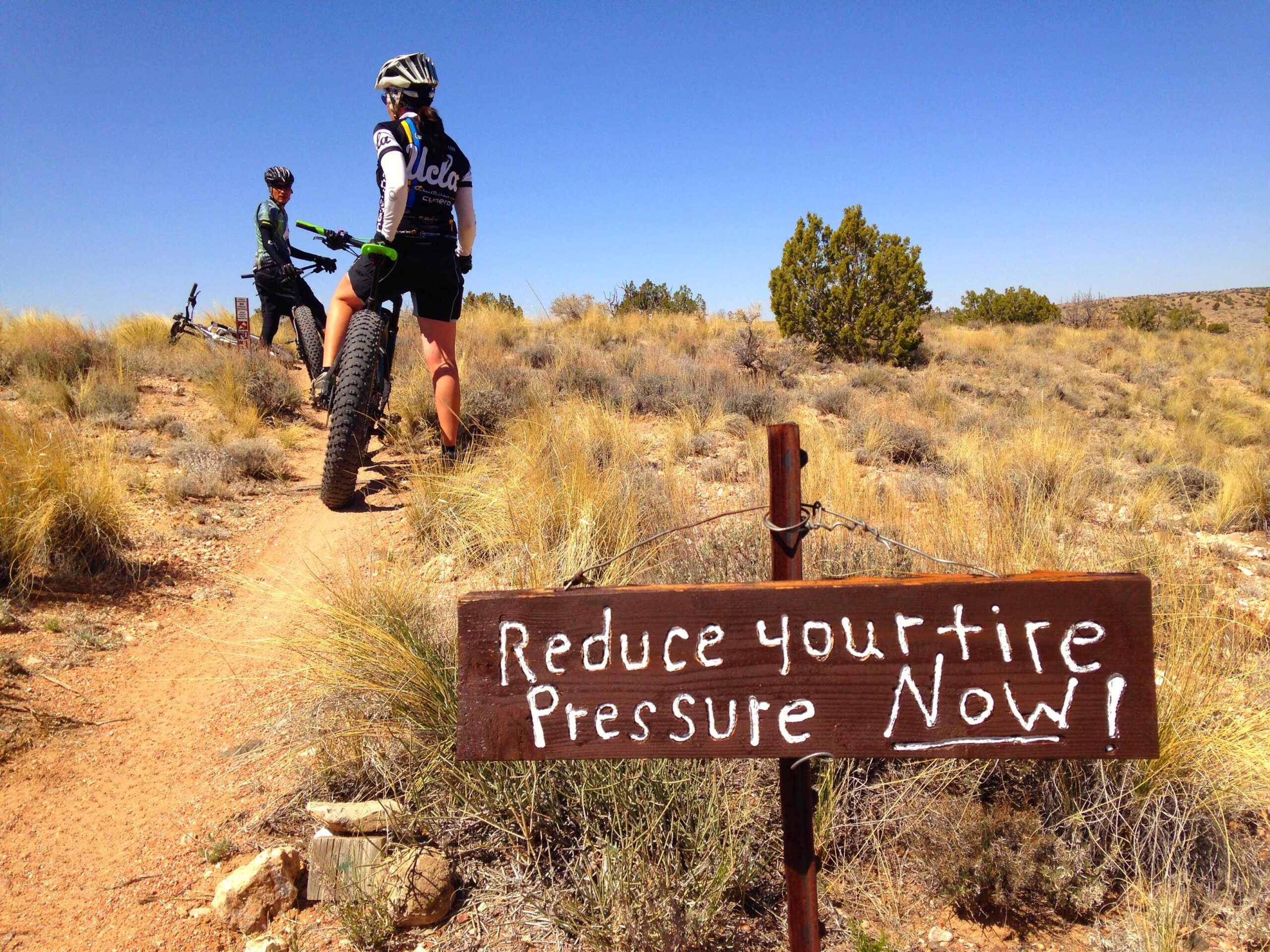 Two mountain bikers pause on a dirt trail surrounded by tall grass and shrubs. A wooden sign in the foreground reads, "Reduce your tire pressure NOW!" against a backdrop of clear blue sky. The bikers, one wearing a UCLA jersey, are positioned beside their bikes, engaged in discussion. Parkway Fatbike trail mountain bike trail.