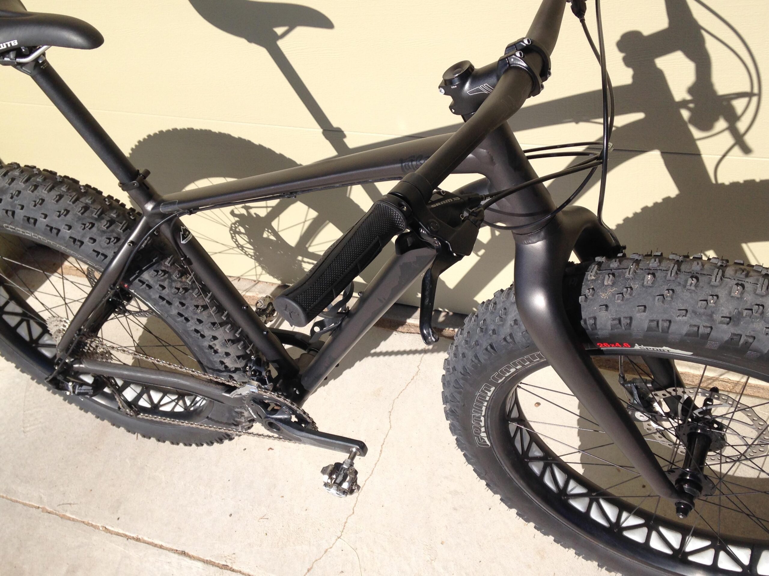 A close-up image of a black fat bike, showcasing its thick tires, sleek frame, and intricate design details, with a shadow cast on a light-colored wall in the background.