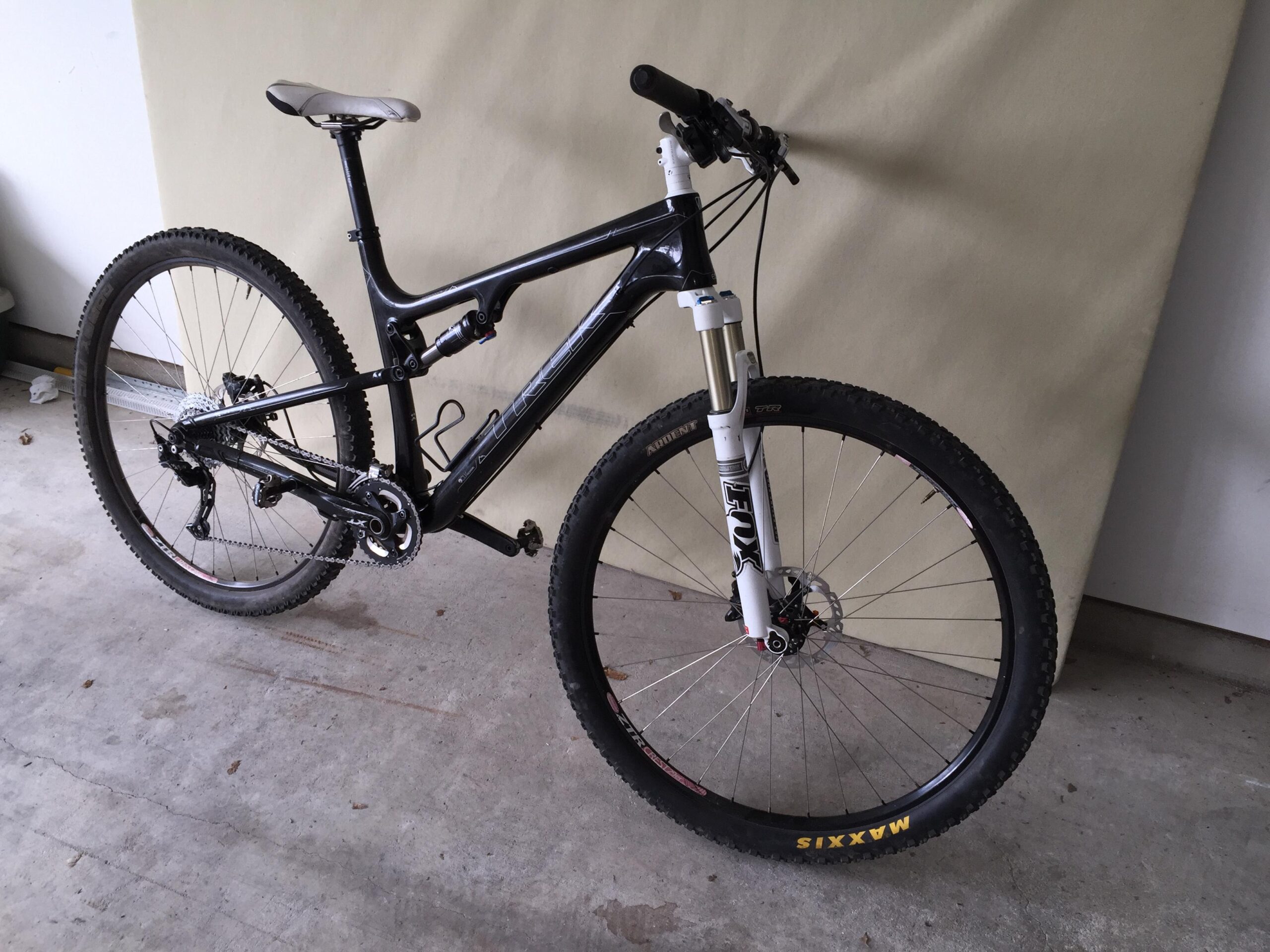 Trek Superfly 100 AL: Mountain bike with a black frame and white front suspension fork, resting on a concrete floor against a neutral-colored backdrop. The bike features thick tires and a modern gear system.