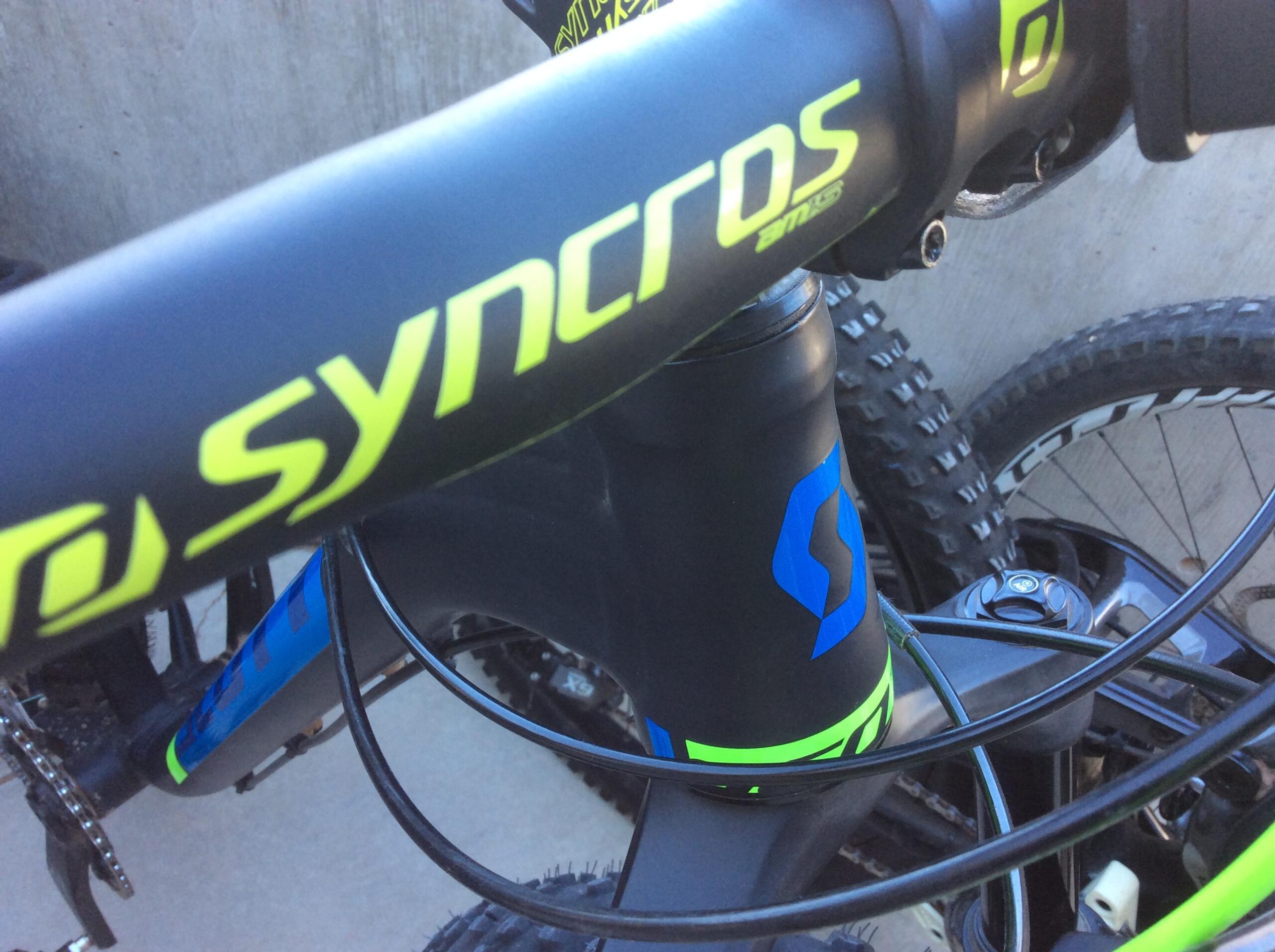 Close-up view of a mountain bike's handlebars and frame, featuring components with green and blue accents, including the SyncrOS logo and cable housing. The background shows part of the bike's wheel and tire.