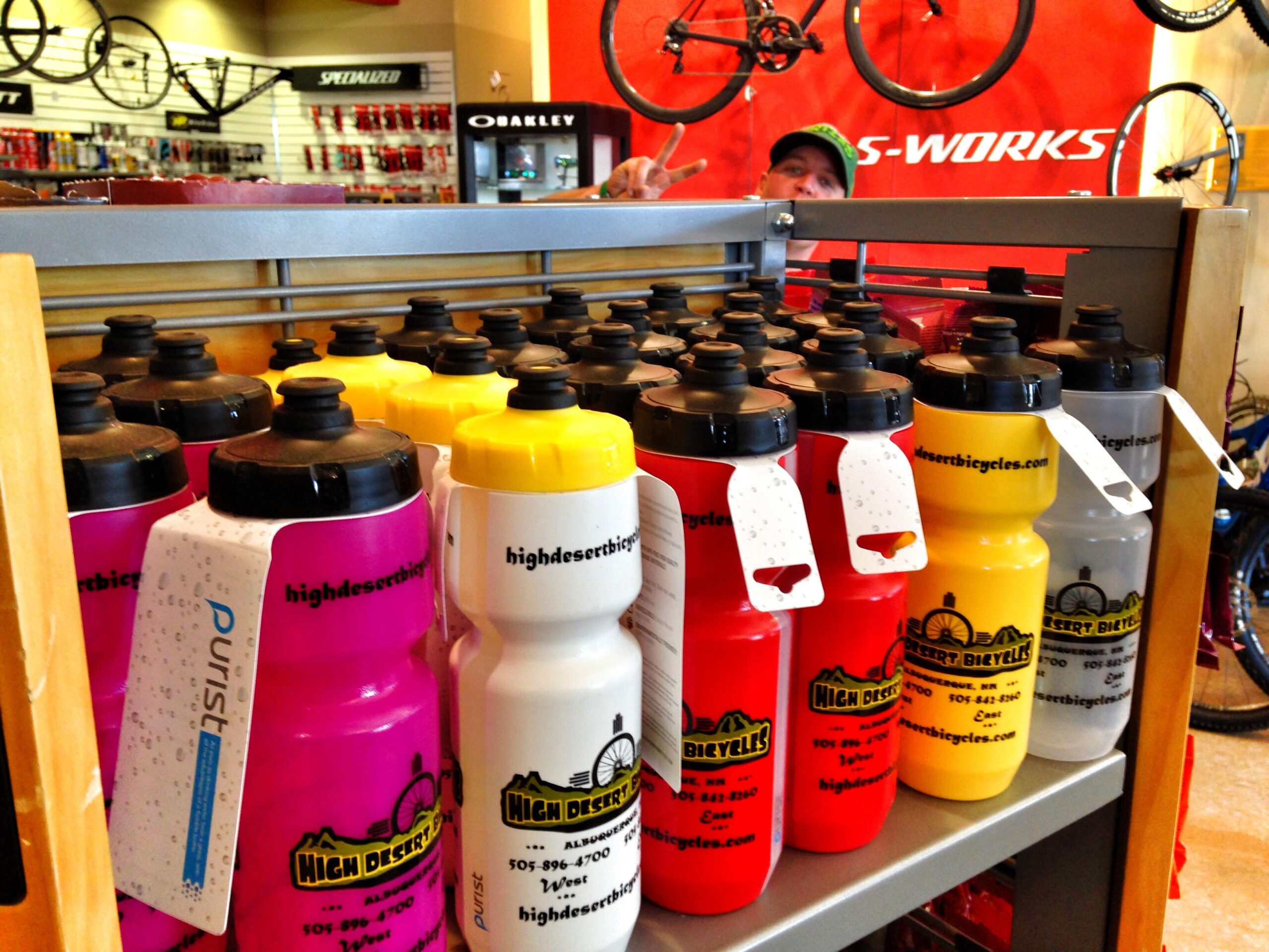 A display of colorful water bottles in a bicycle shop, featuring various colors including pink, red, yellow, and white. The bottles are labeled with the brand "High Desert Bicycles" and include tags with details. Bicycles and bicycle accessories can be seen in the background.