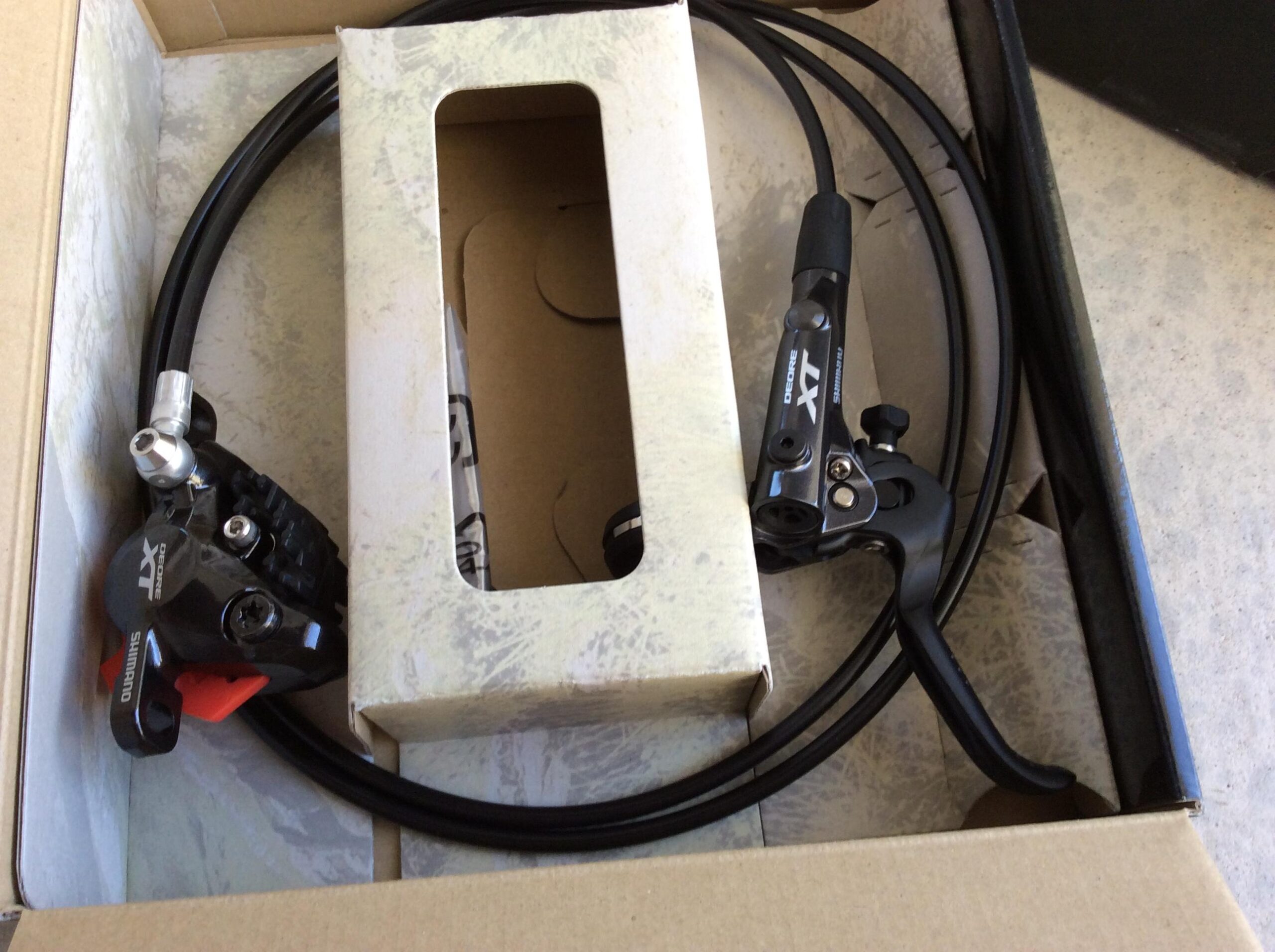 A Shimano XT disc brake system with brake levers and hoses, packaged in a cardboard box with a partially opened lid, showcasing the components inside.