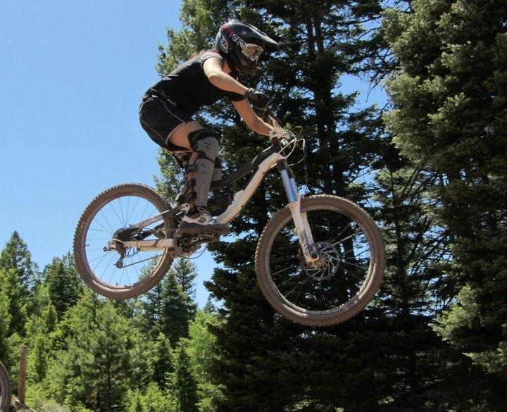 A mountain biker performing a jump over a dirt ramp, wearing protective gear and a helmet, surrounded by tall green trees under a clear blue sky. Angel Fire Bike Park mountain bike trail.