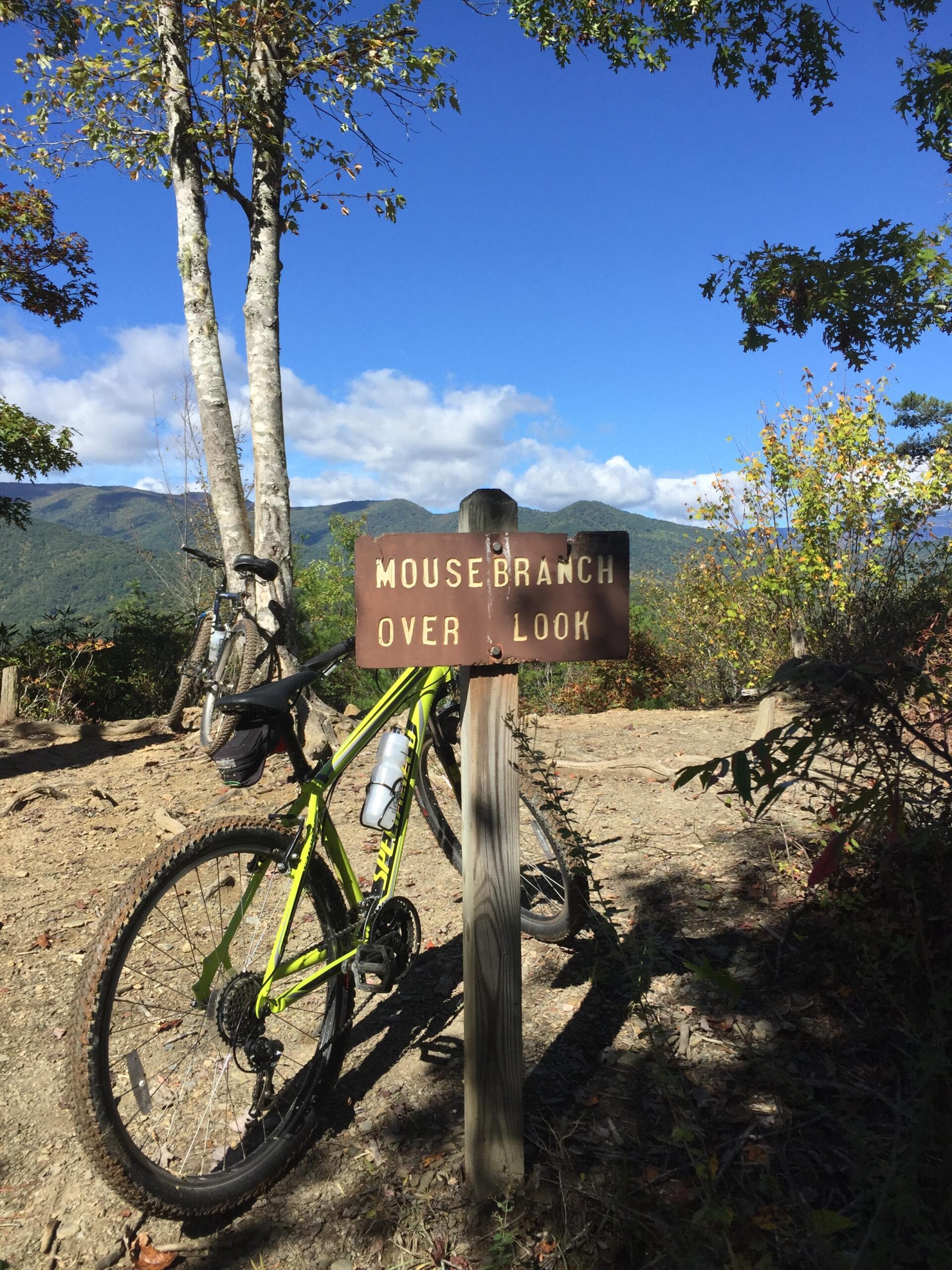 A green mountain bike parked next to a wooden sign that reads "Mouse Branch Overlook," with a scenic view of mountains and a clear blue sky in the background. Tsali Recreation Area mountain bike trail.