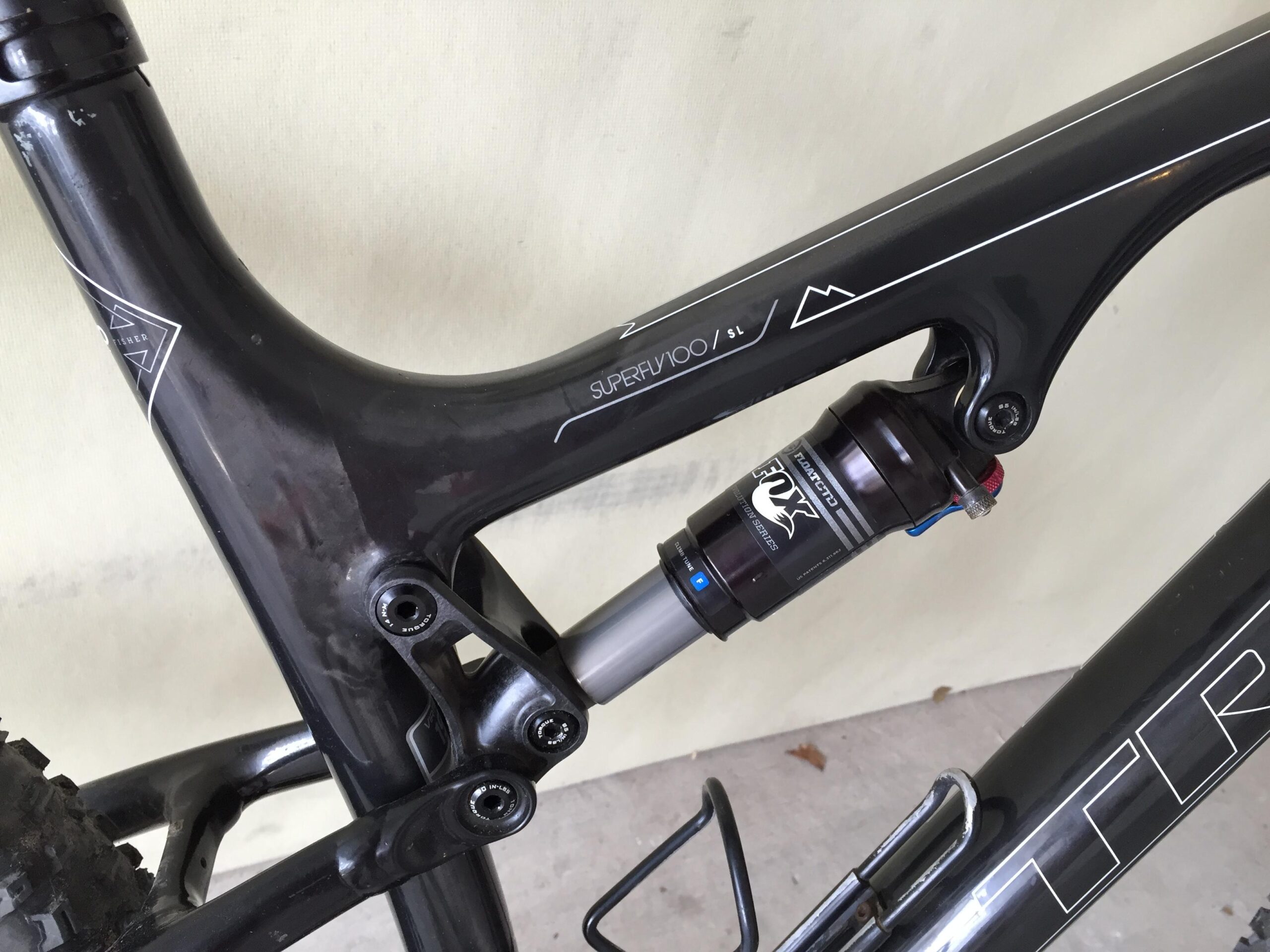 Trek Superfly 100 AL: Close-up view of a bicycle frame showcasing the rear suspension system, with visible components including the shock absorber and frame design details, all in a black finish. The background is a neutral color, helping to highlight the bike's features.