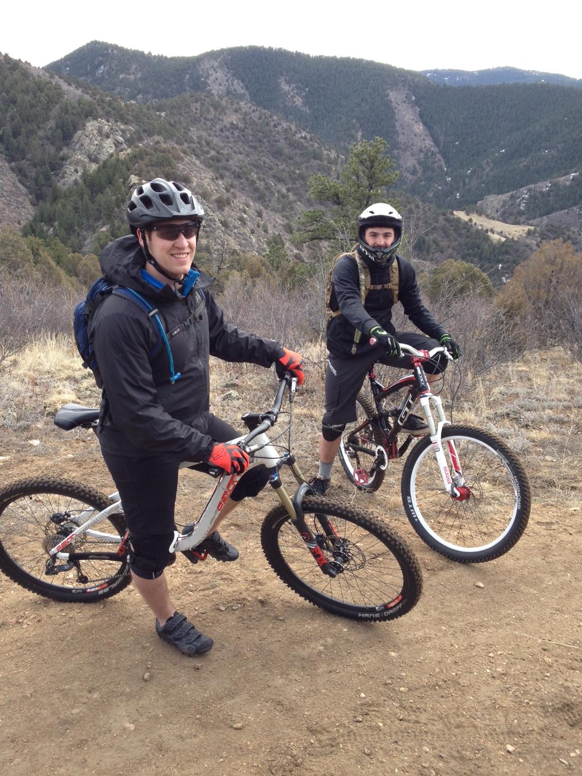 Giant Reign 2: Two mountain bikers standing on a dirt trail with a scenic mountainous background. One biker is wearing a black helmet and jacket, smiling and resting a foot on his bike, while the other wears a white helmet and dark gear, holding onto his bike. The landscape features rolling hills and trees, indicating a natural outdoor setting.