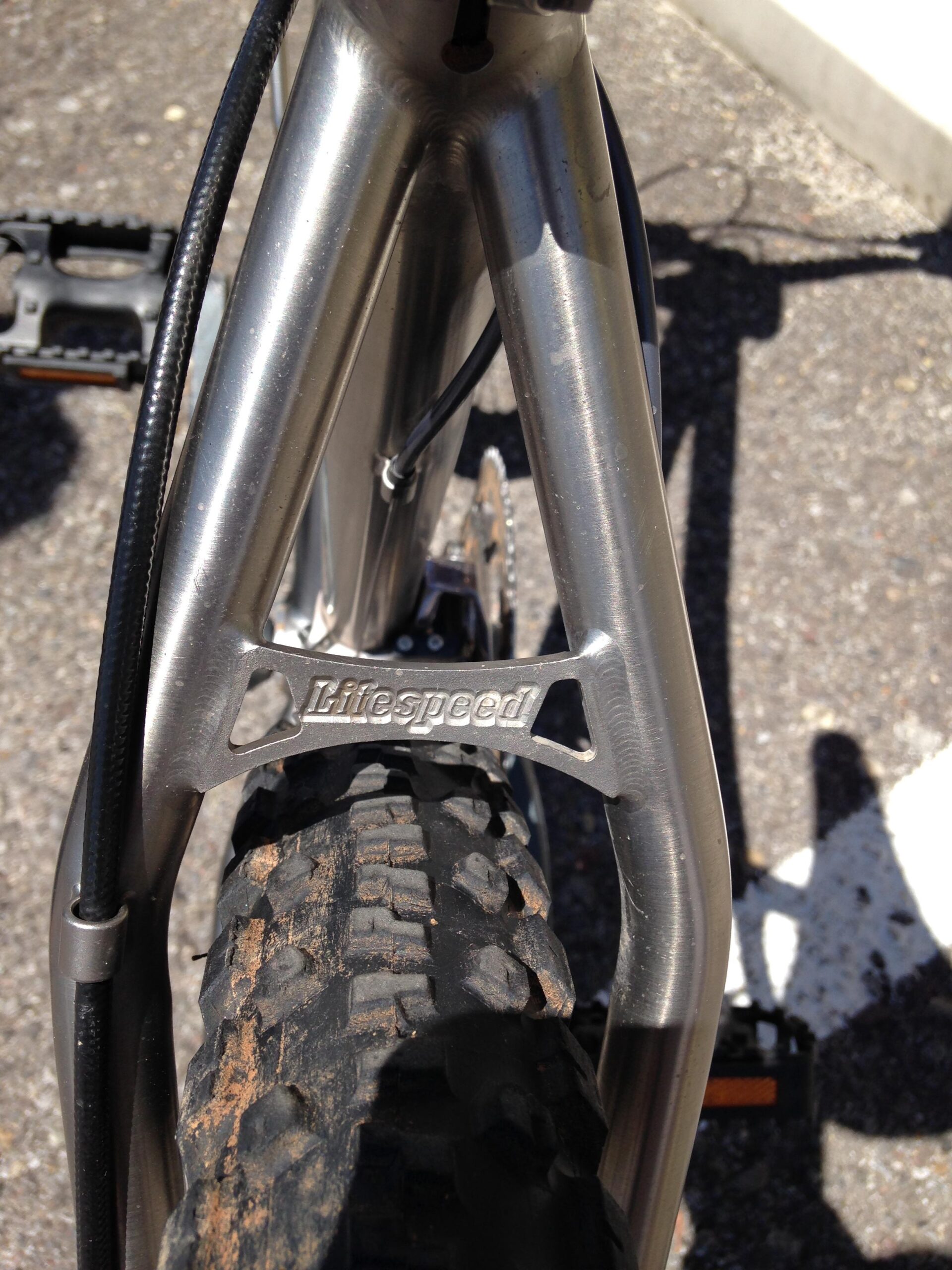 Close-up view of a bicycle frame featuring a metallic finish, with a visible "Litespeed" logo on the rear triangle. The image also shows part of a textured tire and a pedal in the background. The surface beneath is gravel or pavement. Parkway Fatbike trail mountain bike trail.