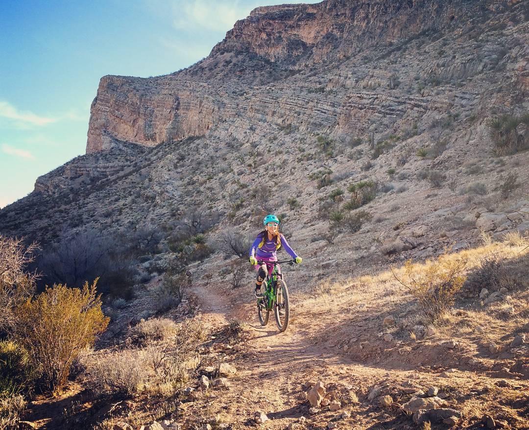 A mountain biker rides along a rocky trail in a desert landscape, with steep, rugged cliffs in the background. The cyclist wears a blue helmet and a purple long-sleeve shirt while navigating the path surrounded by sparse vegetation. The sky above is clear with hints of clouds. Cottonwood Valley North mountain bike trail.