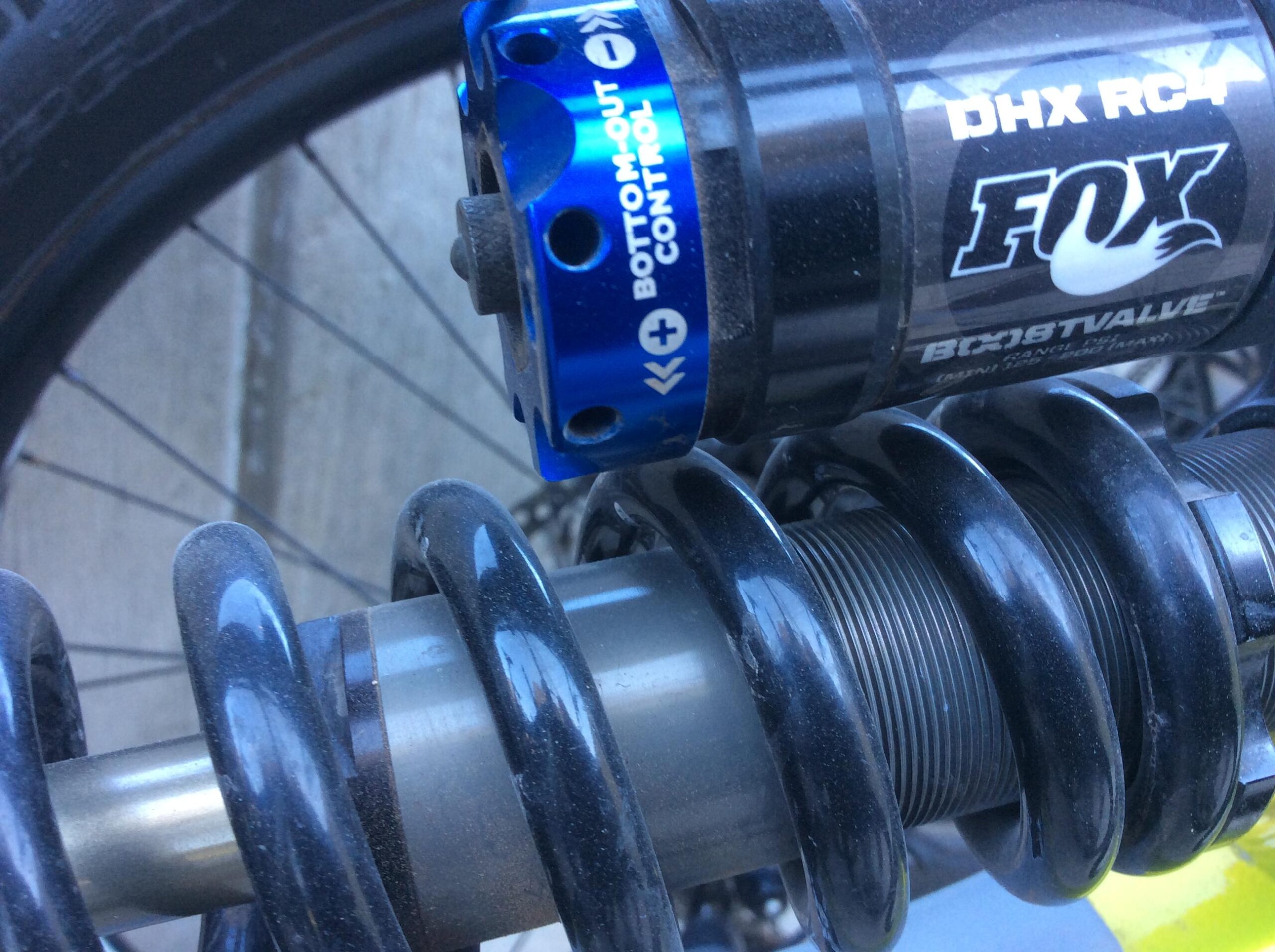 Close-up view of a FOX DHX RC4 mountain bike shock absorber, featuring a black coil spring and a blue bottom-out control knob. The shock shows some signs of wear and is positioned near the rear wheel of a bike.
