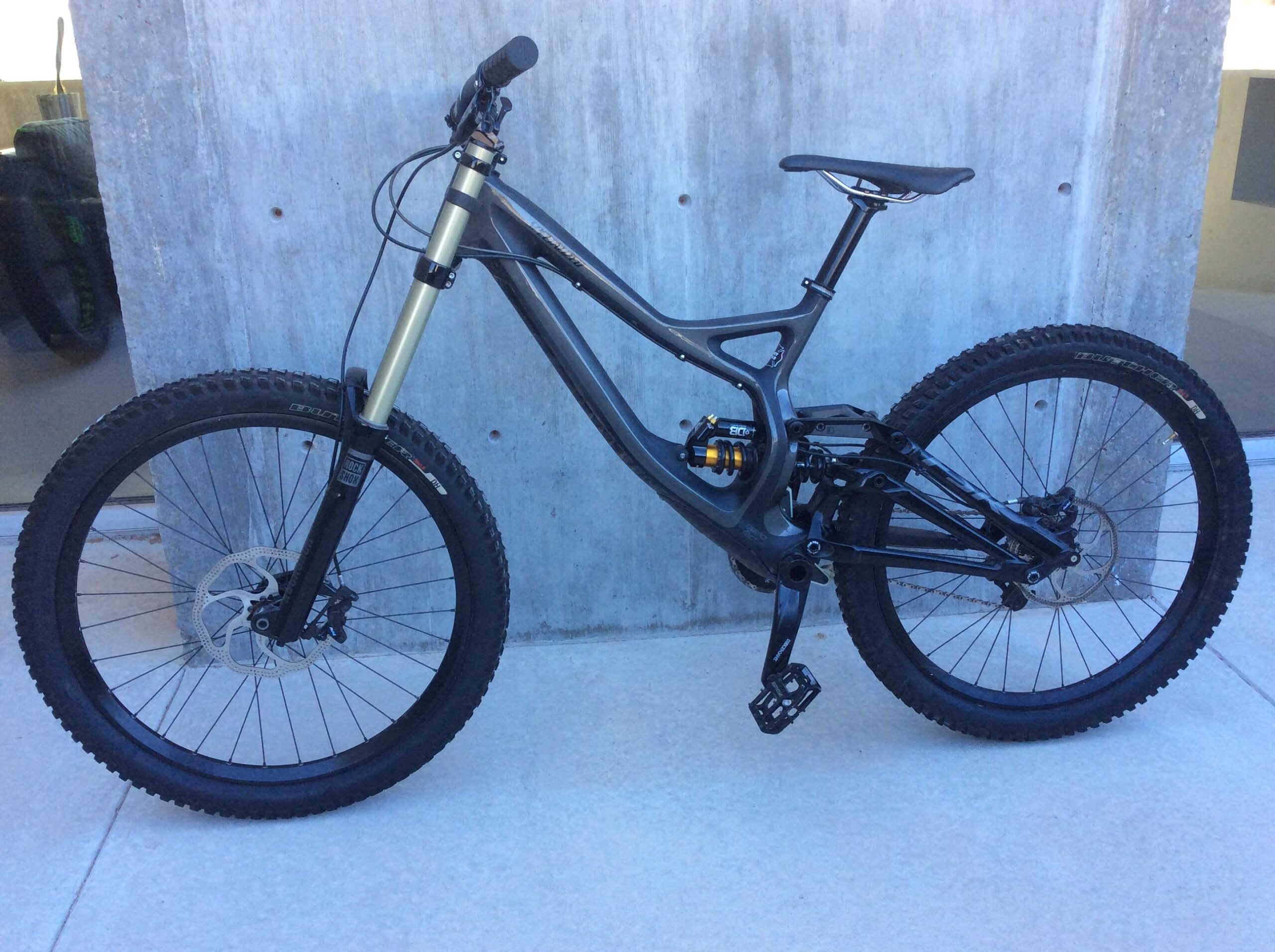 Specialized S-Works Demo 8 Carbon: Image of a modern mountain bike with a sleek design, featuring a black and gray frame, large knobby tires, and a dual suspension system. The bike is positioned against a concrete wall on a flat surface, highlighting its robust build and off-road capabilities. The front fork has a gold-colored shock absorber, and the bike has components designed for high performance and durability.