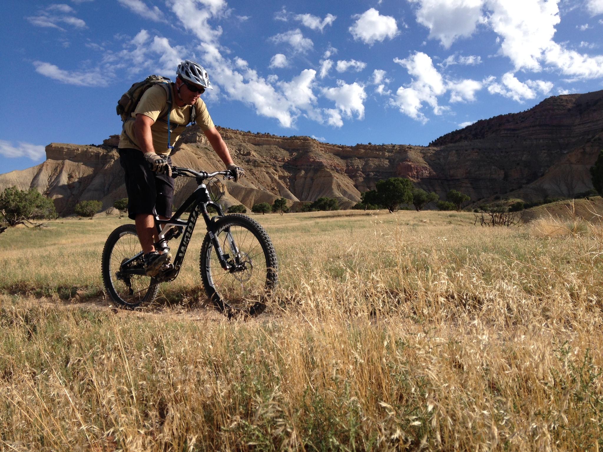 A mountain biker riding through a grassy field with hills and blue skies in the background. The cyclist is wearing a helmet and a backpack, focused on the trail ahead. 18 Road Trails / North Fruita Desert mountain bike trail.