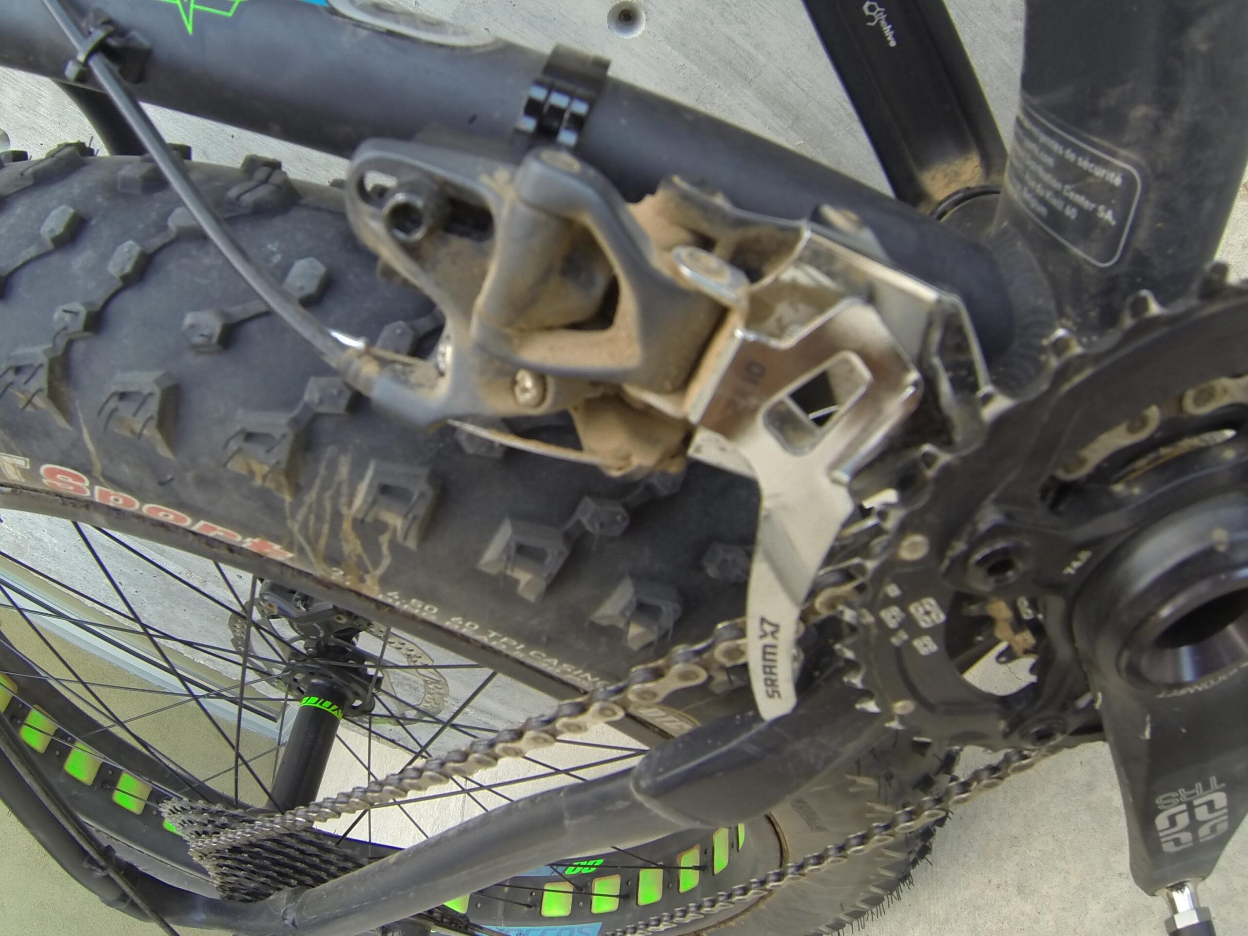 Scott Big Ed: Image showing a close-up view of a mountain bike's drivetrain, including the derailleur, chain, and gear system, with visible dirt and wear indicating usage. Green accents on the bike frame and wheel.