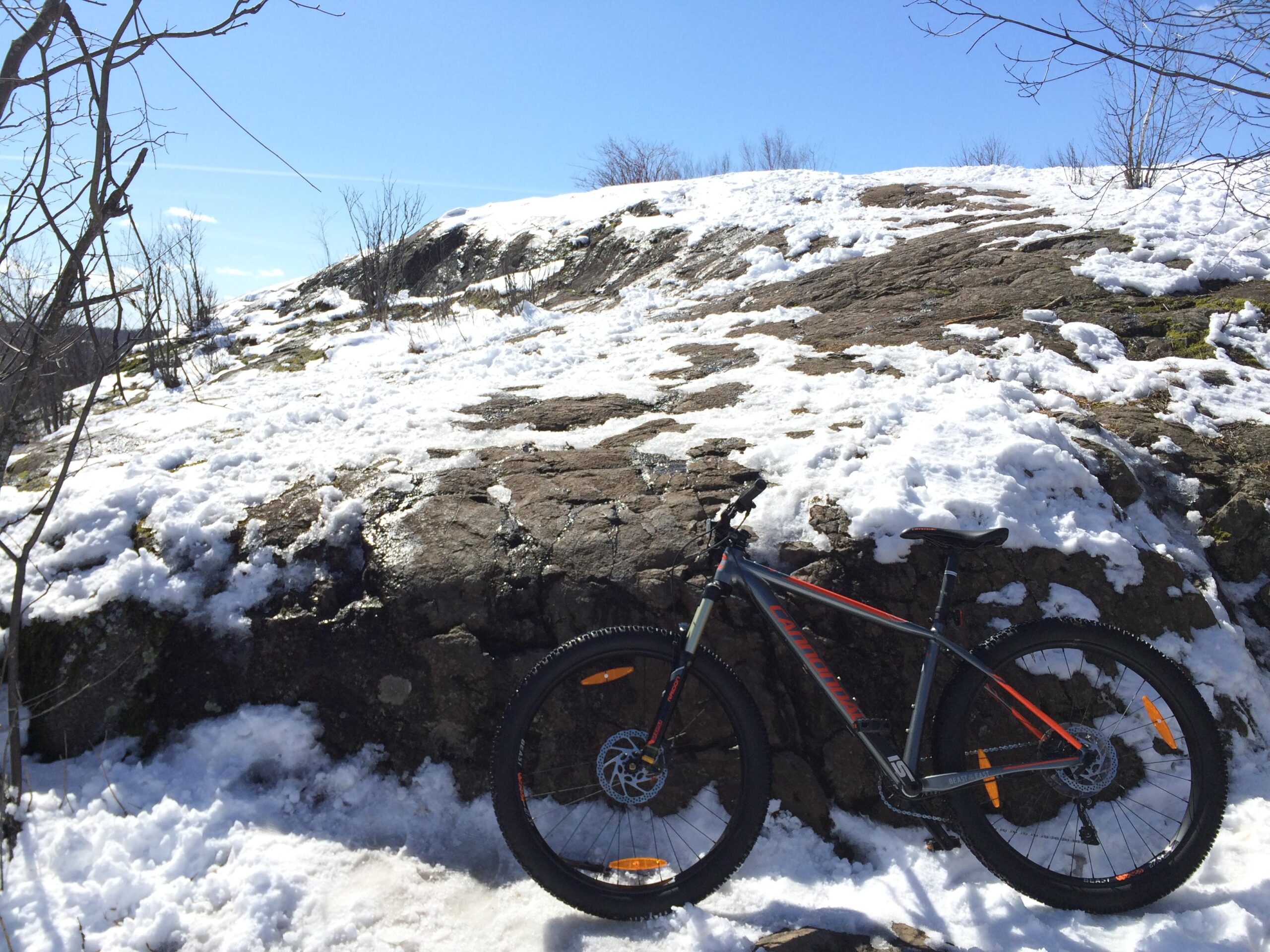 A mountain bike resting against a rocky surface partially covered in snow, under a clear blue sky. Hartley Park mountain bike trail.