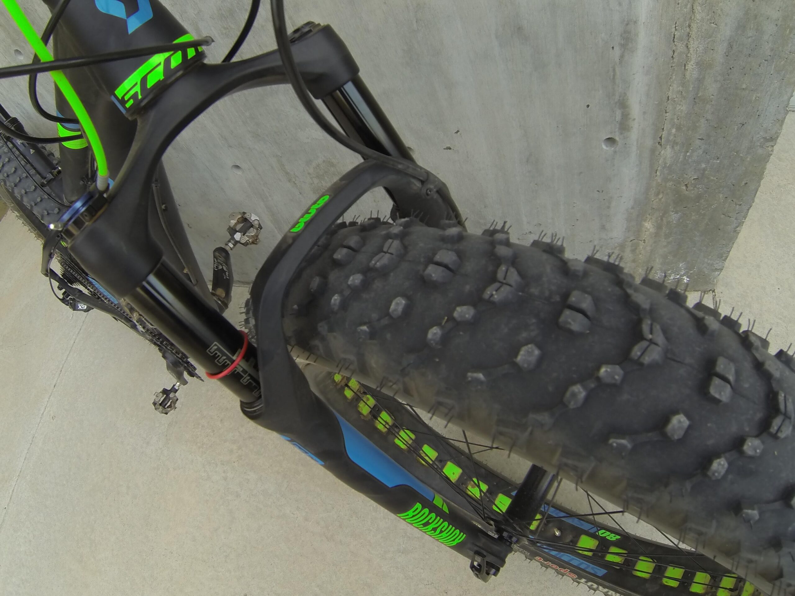 Scott Big Ed: Close-up view of a mountain bike's front fork and wheel, showcasing the textured tread of the tire against a concrete background. The bike features a black and green color scheme with visible components like the suspension and pedals.