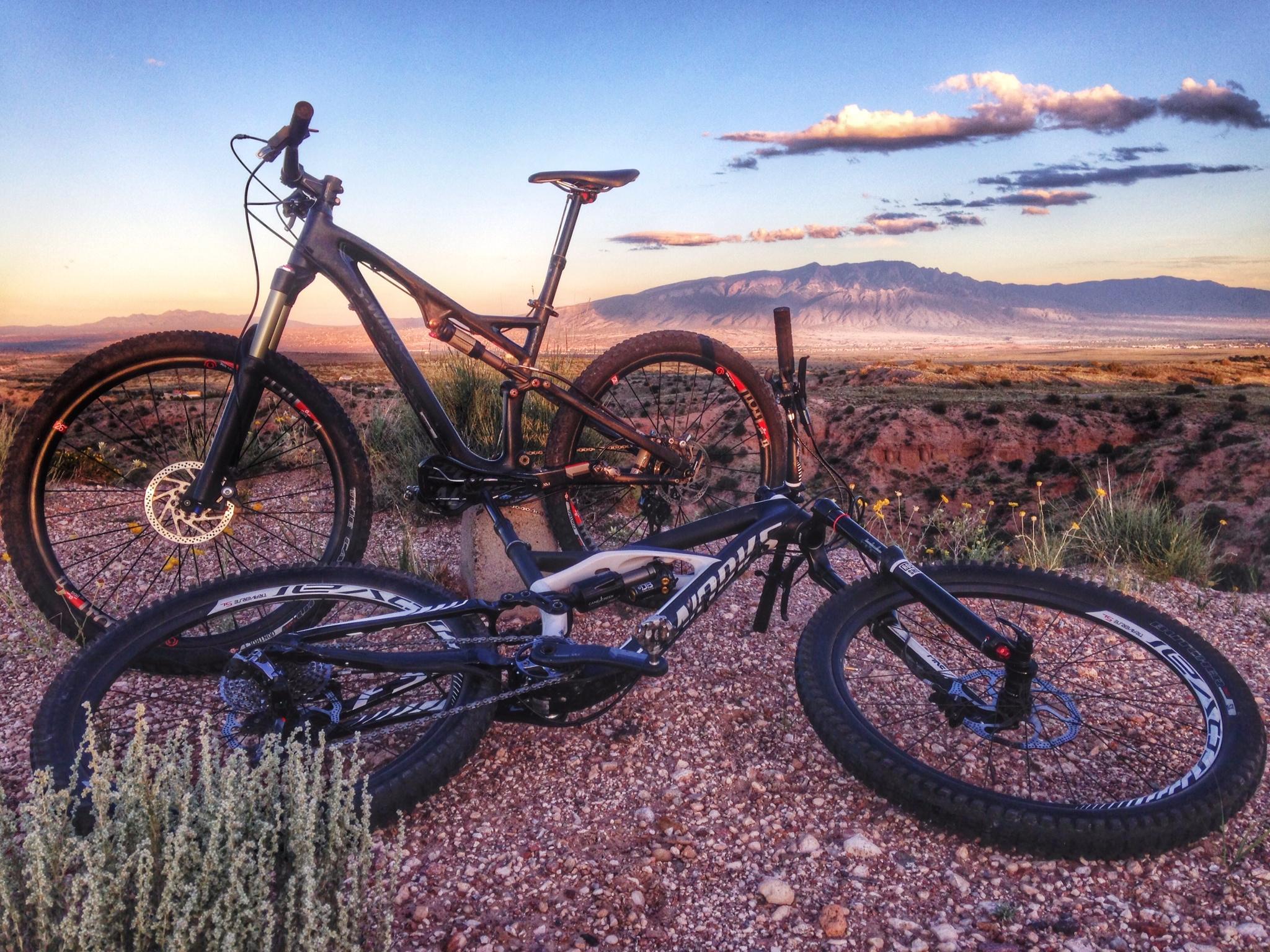 Specialized Stumpjumper FSR Expert: Two mountain bikes are positioned on a gravelly terrain with a scenic view of mountains and a colorful sky during sunset. The bikes feature distinct designs, one standing upright while the other is lying on its side, surrounded by sparse vegetation. The landscape showcases a dramatic mountainous backdrop under a sky filled with clouds painted in warm hues.