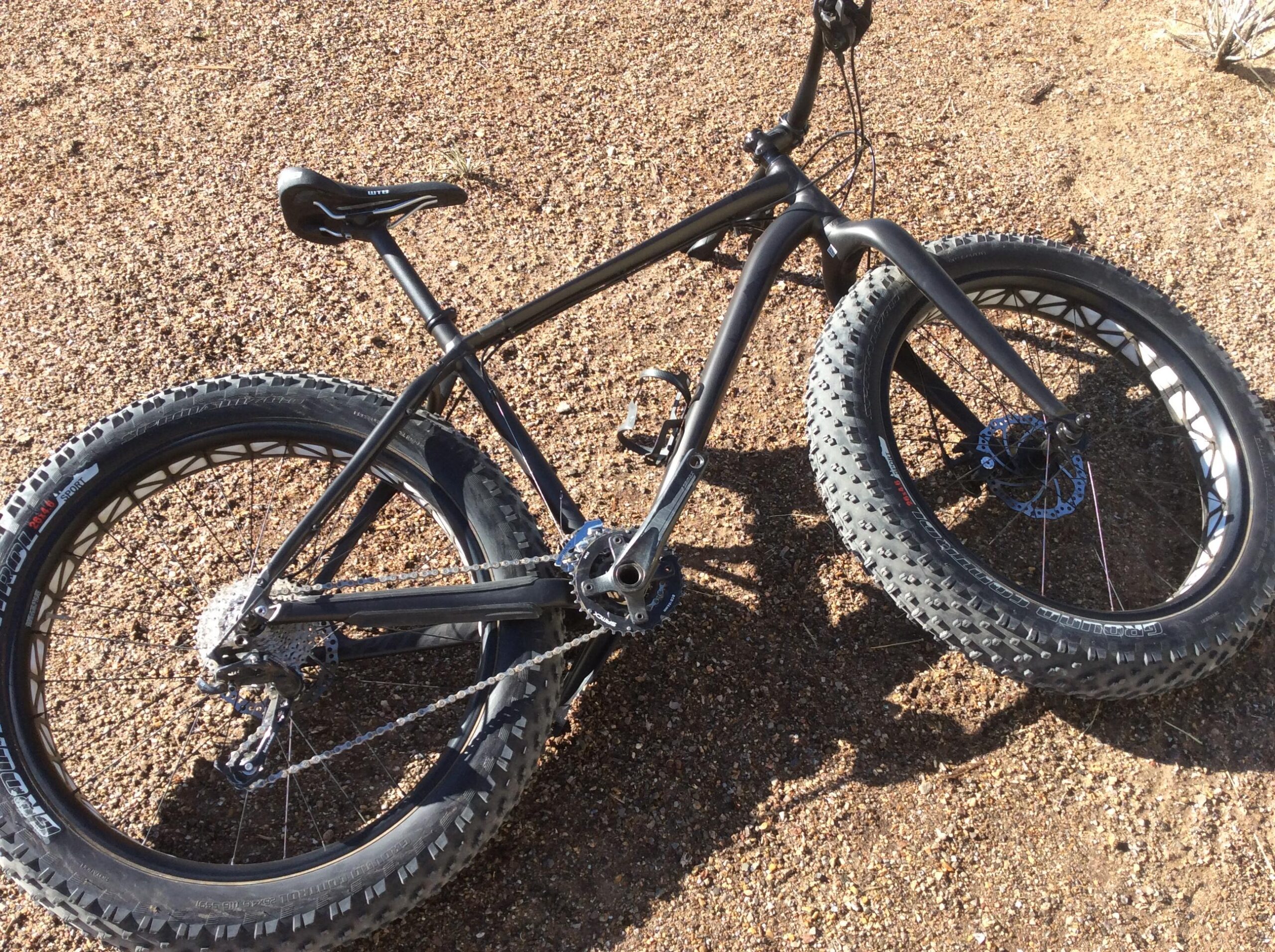 Specialized Fatboy: A black fat bike lying on a gravel surface, showcasing large, knobby tires and a minimalist frame design. The bike's chain and gears are visible, emphasizing its rugged build suitable for off-road cycling.
