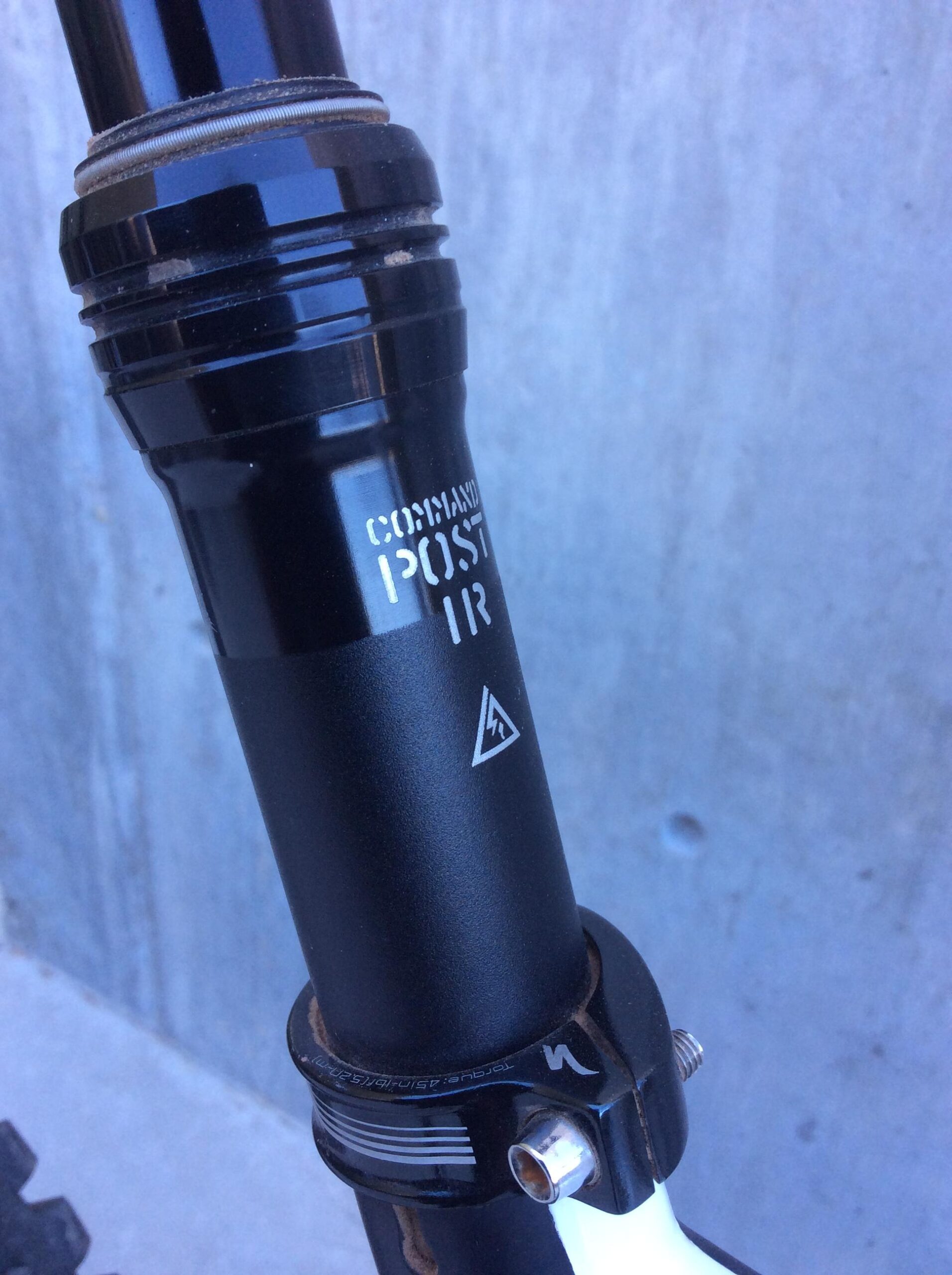 Close-up image of a black bike dropper post with the label "COMMAND POST IR" visible. The post is attached to a bike frame featuring a circular clamp and a threaded bolt. A textured, concrete background is partially visible.