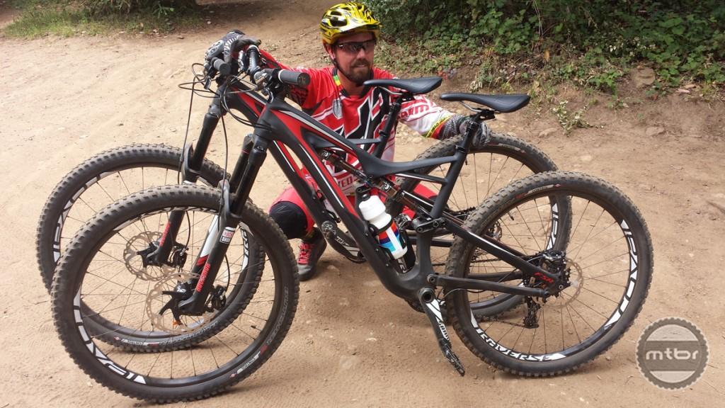 A mountain biker sitting on the ground next to two bicycles, showcasing their unique designs. The biker is wearing protective gear and a helmet, with a dirt path and greenery in the background, indicating an outdoor cycling environment.
