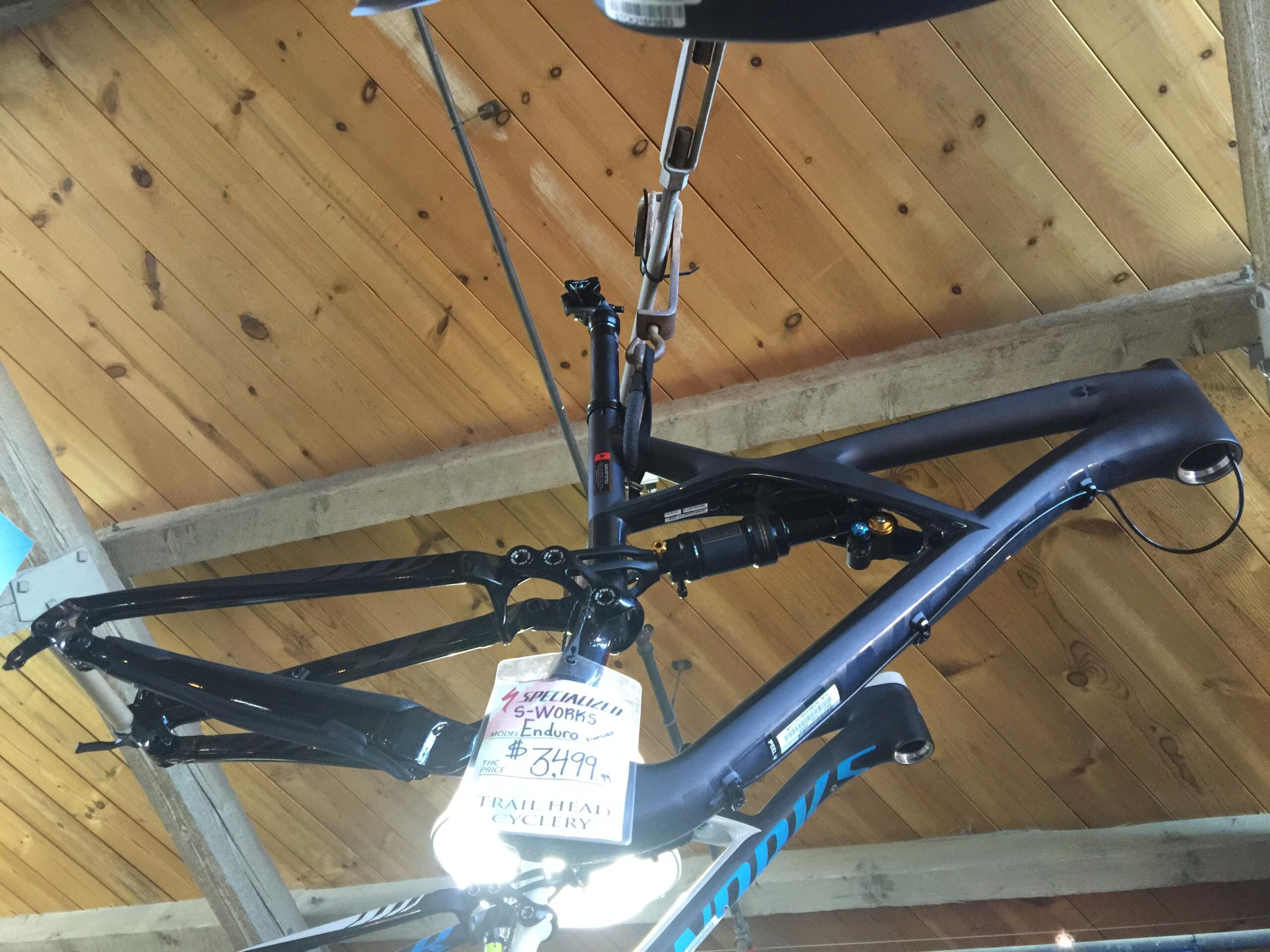 A close-up view of a Specialized S-Works Enduro bike frame hanging from the ceiling of a bike shop. The frame is predominantly black with some blue accents, and a price tag reading $3,499 is attached. The wooden ceiling and beams of the shop are visible in the background.