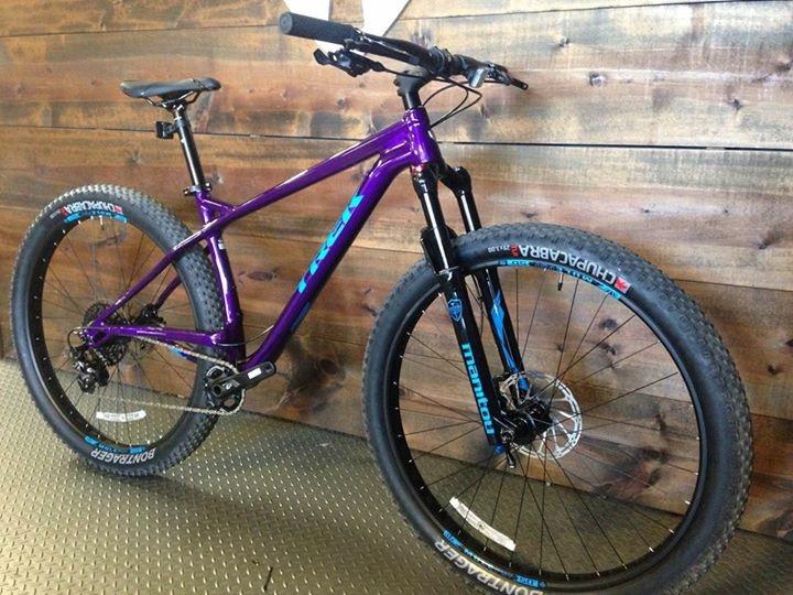 Trek Stache: A purple mountain bike with a lightweight frame and thick tires, positioned against a wooden wall. The bike features prominent suspension forks, disc brakes, and stylized branding on the tires, emphasizing its off-road capabilities.