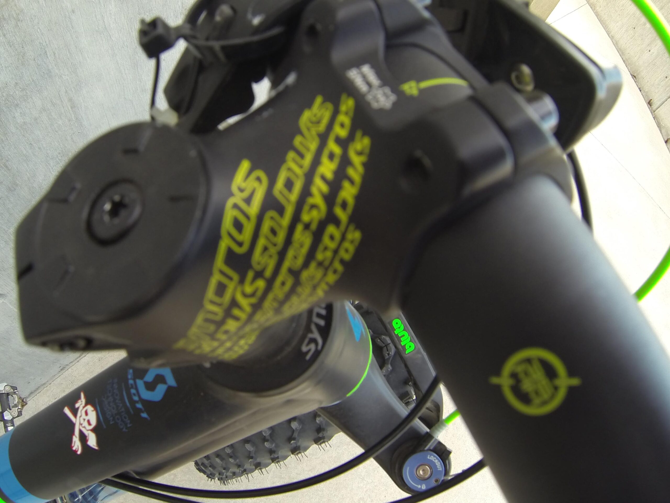 Scott Big Ed: Close-up image of a mountain bike's handlebar and stem, featuring black components with vibrant green and yellow accents. The bike's branding and design details are visible, highlighting the intricate mechanics and style of the equipment. The background is a blurred concrete surface, suggesting an outdoor setting.