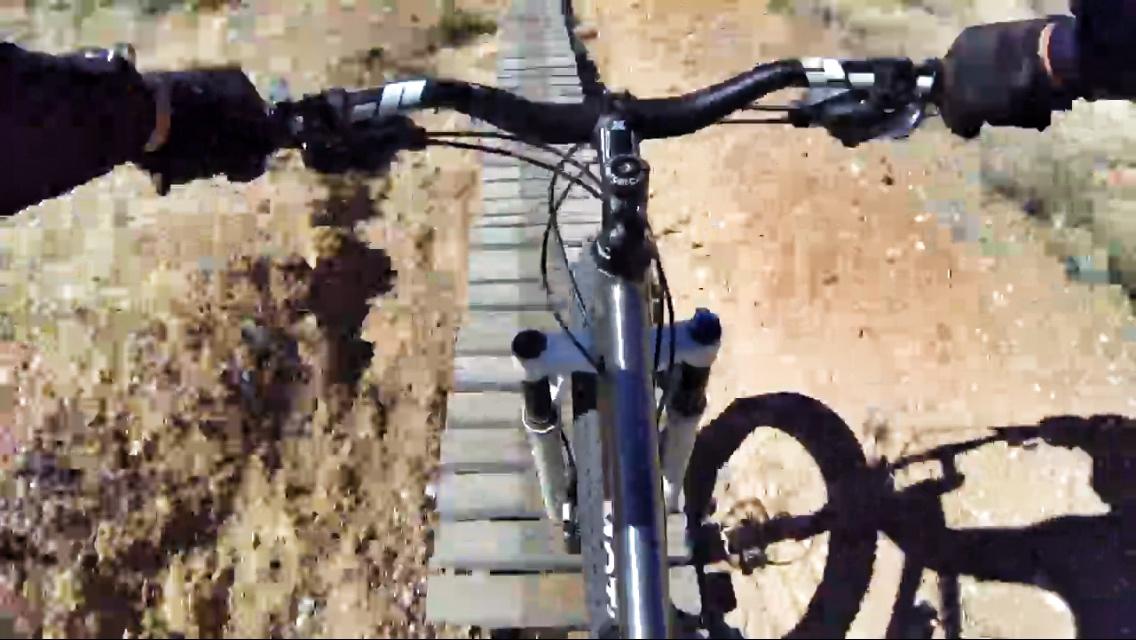 A first-person view of a mountain bike navigating a narrow wooden plank path over rough terrain. The handlebars are visible in the foreground, with dirt and dry vegetation on either side of the path. Goat FatBike Trail mountain bike trail.