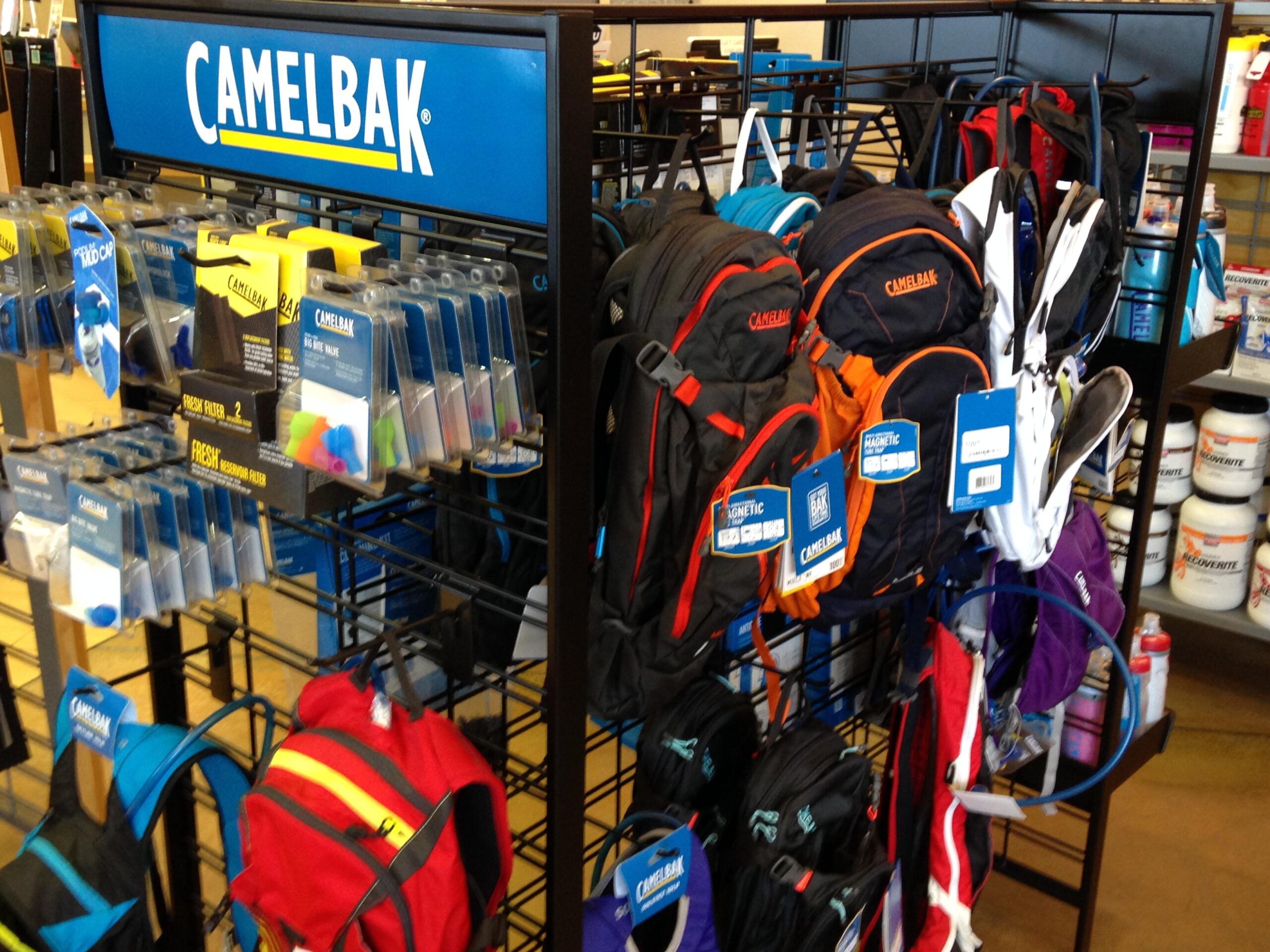 Display of CamelBak products, including various hydration packs and accessories, arranged on a black metal rack. The top features a blue sign with the CamelBak logo, and there are tags attached to several bags highlighting their features. The background includes additional items such as bottles and containers.