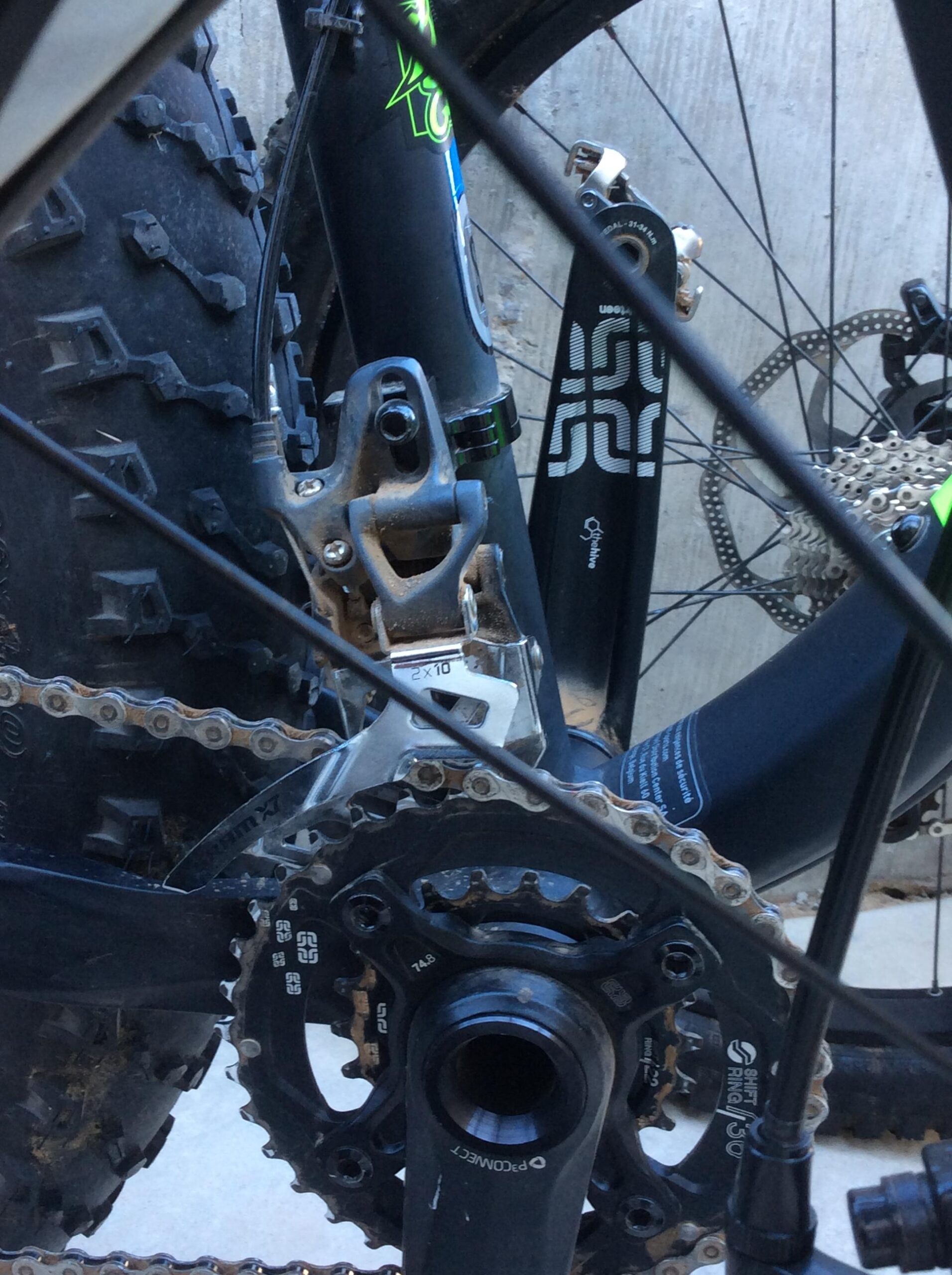 Close-up of a bicycle drivetrain system, featuring a gear shifter, chainrings, and a chain, with a textured tire partially visible in the background. The bike components display dirt, indicating recent use.