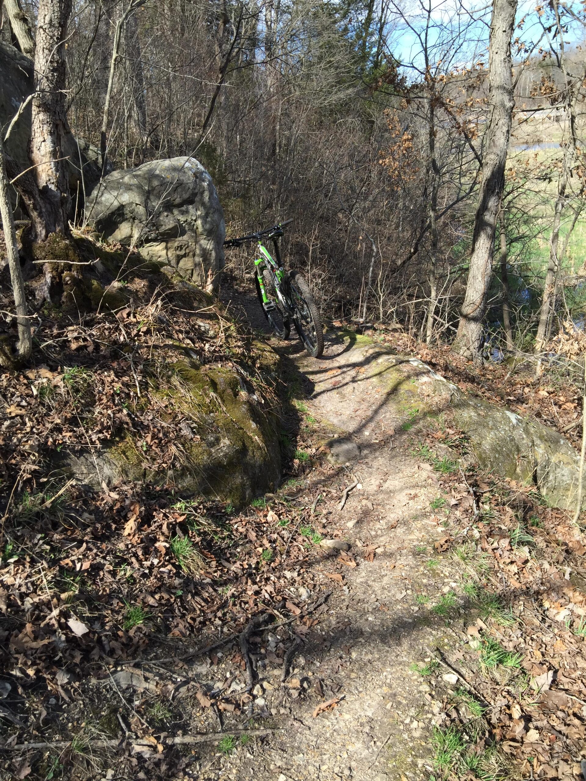 A narrow dirt trail surrounded by trees and rocky terrain, with a mountain bike leaning against a large rock. The scene is set in a natural outdoor environment with fallen leaves and patches of green grass. Binder Lake mountain bike trail.