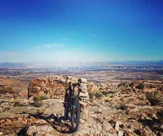 A mountain bike resting on rocky terrain, with a cairn stacked nearby. In the background, a panoramic view of a valley and distant city skyline under a clear blue sky. Cowboy Trails mountain bike trail.