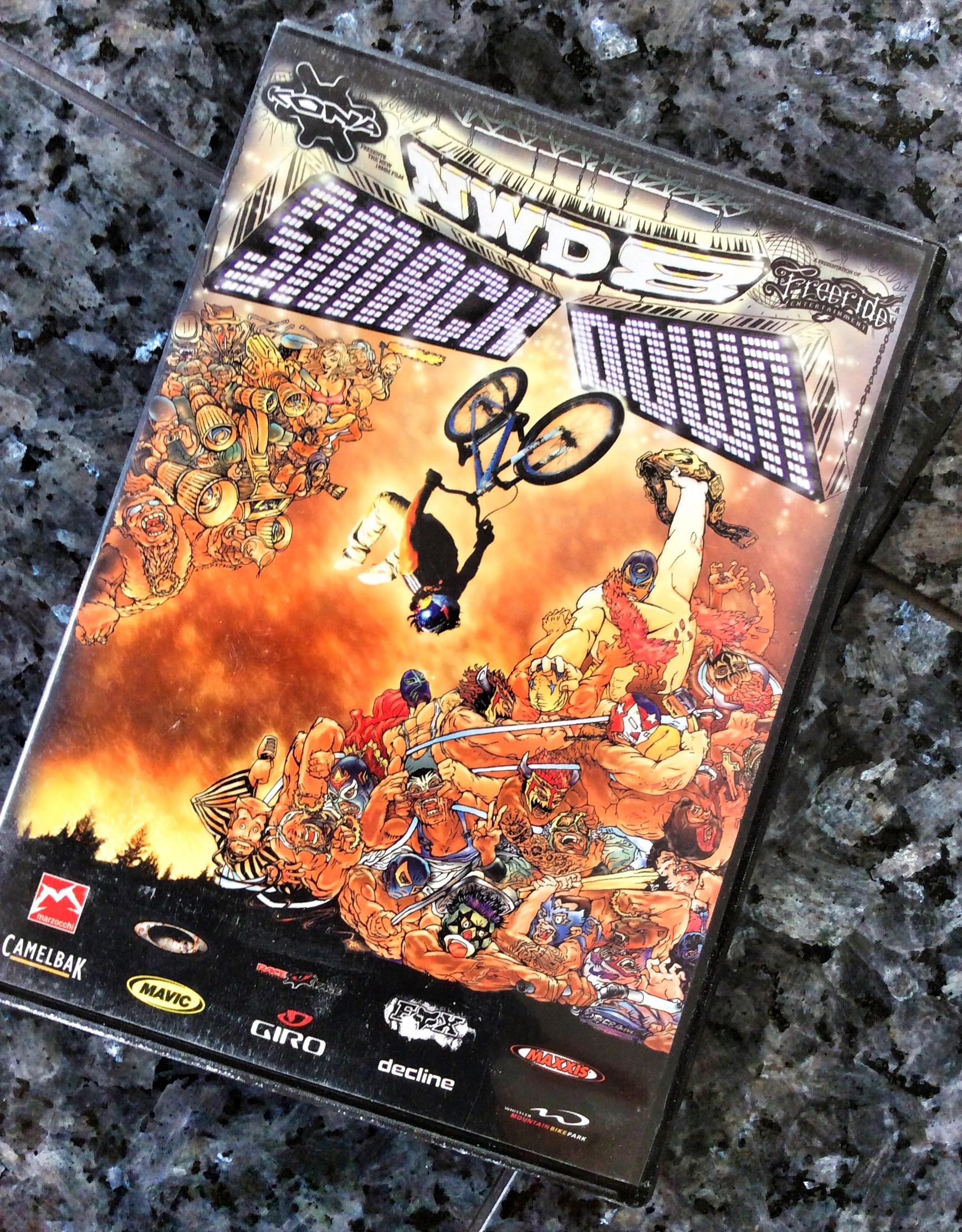 Image of a video game cover titled "NWDS Smack Down." The cover features vibrant, comic-style art with a central figure performing a trick on a bicycle, surrounded by a chaotic scene of various characters in dynamic poses. The background is a fiery gradient, enhancing the action-oriented theme. Logos of different brands and sponsors are located at the bottom of the cover.