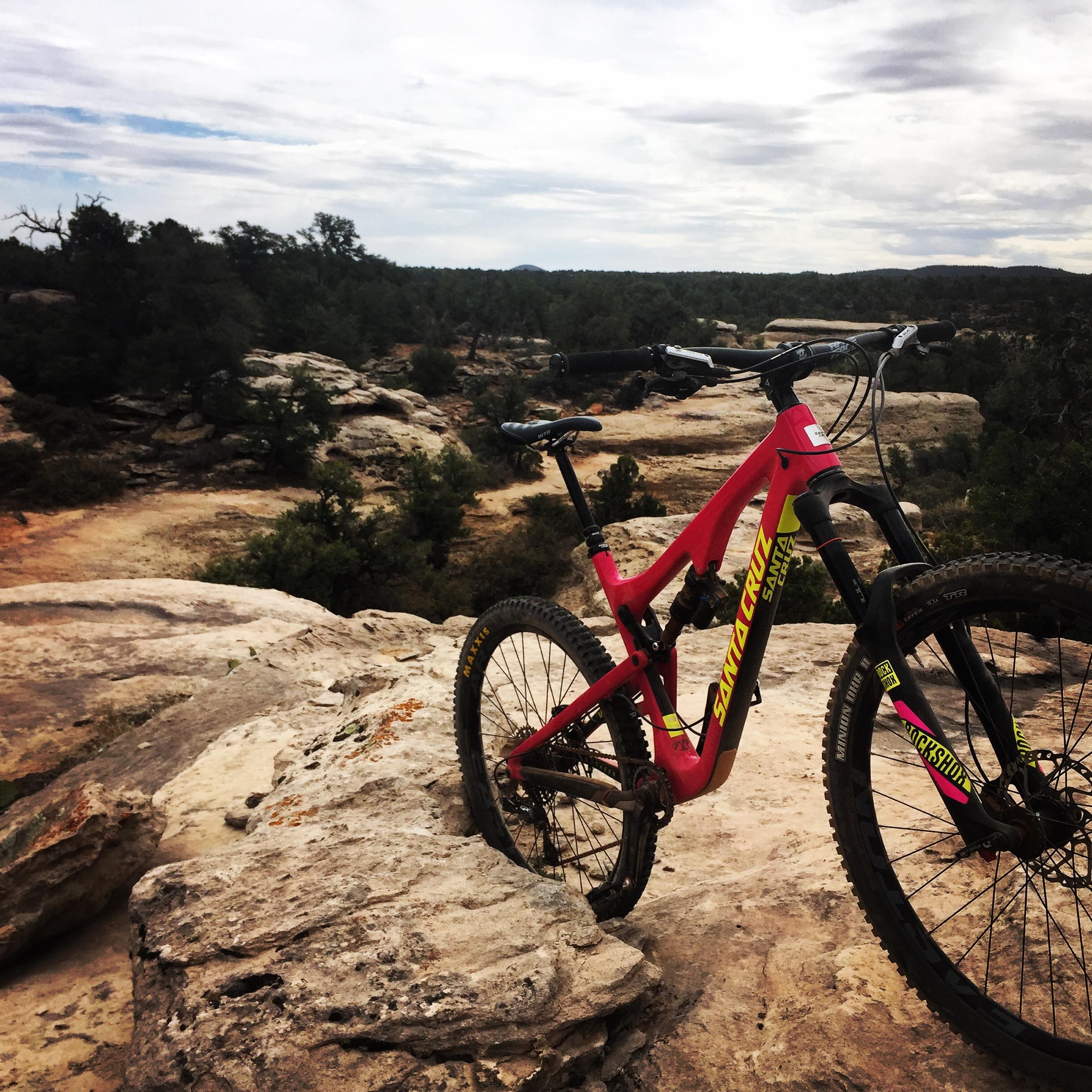 Santa Cruz Bronson: A bright pink mountain bike is positioned on a rocky outcrop, overlooking a rugged landscape with sparse trees and rolling hills under a cloudy sky.
