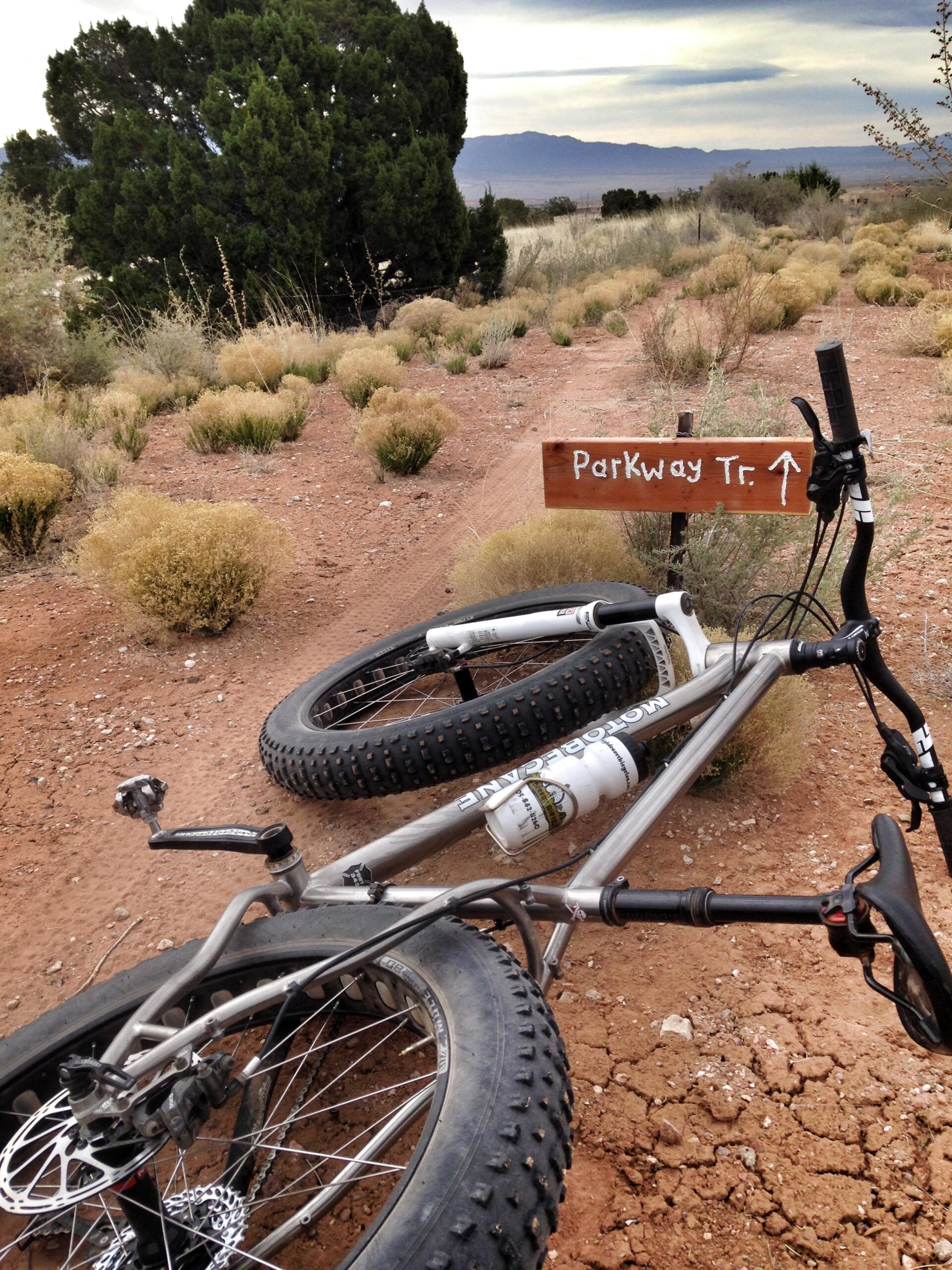 A mountain bike lies on its side in a sandy landscape, with a wooden signpost labeled "Parkway Tr." pointing to a nearby bike trail. Surrounding the bike are sparse shrubs and grasses, with distant mountains visible under a cloudy sky. Parkway Fatbike trail mountain bike trail.