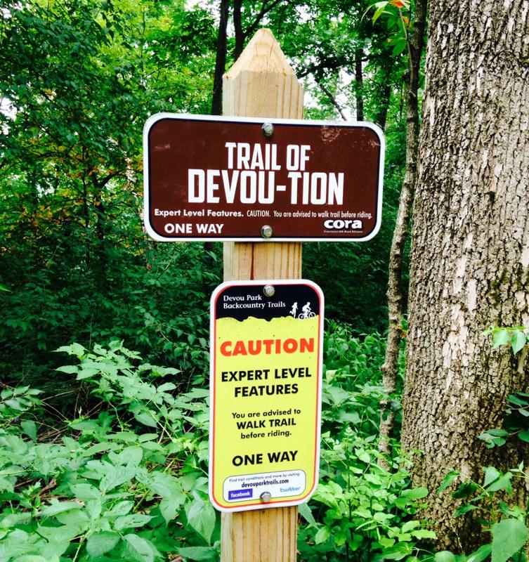 A wooden signpost in a wooded area with two signs: the top sign reads "Trail of Devou-tion" with a note about expert level features and a warning to walk the trail before riding, while the bottom sign emphasizes caution for expert level features and states the trail is one way. Surrounding the signpost are green foliage and trees. Devou Park mountain bike trail.