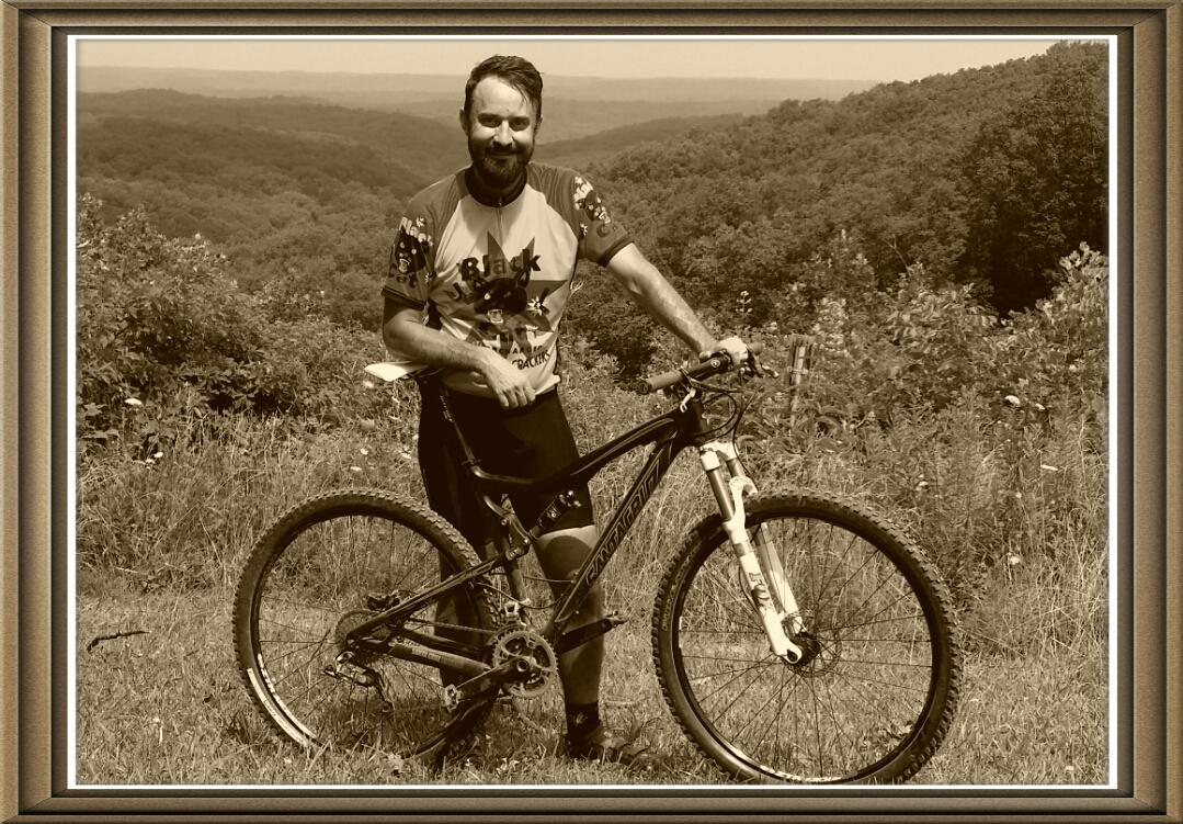 A man stands next to his mountain bike on a grassy hillside, surrounded by a scenic view of rolling hills and trees. He is wearing a colorful cycling jersey and black shorts, smiling as he holds onto the bike. The image has a sepia tone, giving it a vintage feel. Brown County Park mountain bike trail.