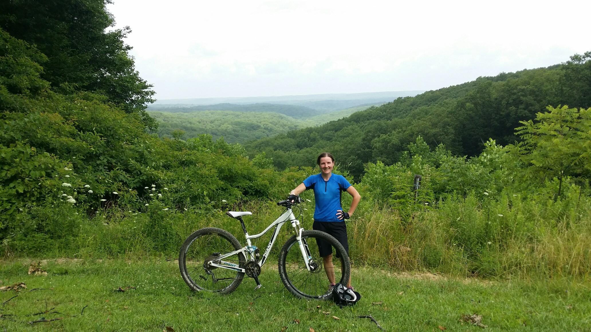 A smiling person stands beside a mountain bike in a grassy area with a scenic view of rolling green hills in the background. The sky is overcast, indicating a cloudy day. The surroundings are lush with trees and plants, creating a natural, outdoor atmosphere. Brown County Park mountain bike trail.