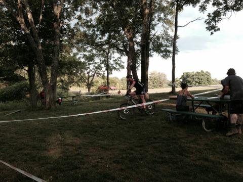 A cyclist navigating a course marked by tape through a grassy area surrounded by trees, with people engaged in various activities, including sitting at picnic tables. The setting appears to be part of an outdoor event. England Idlewild Mountain Biking Park mountain bike trail.